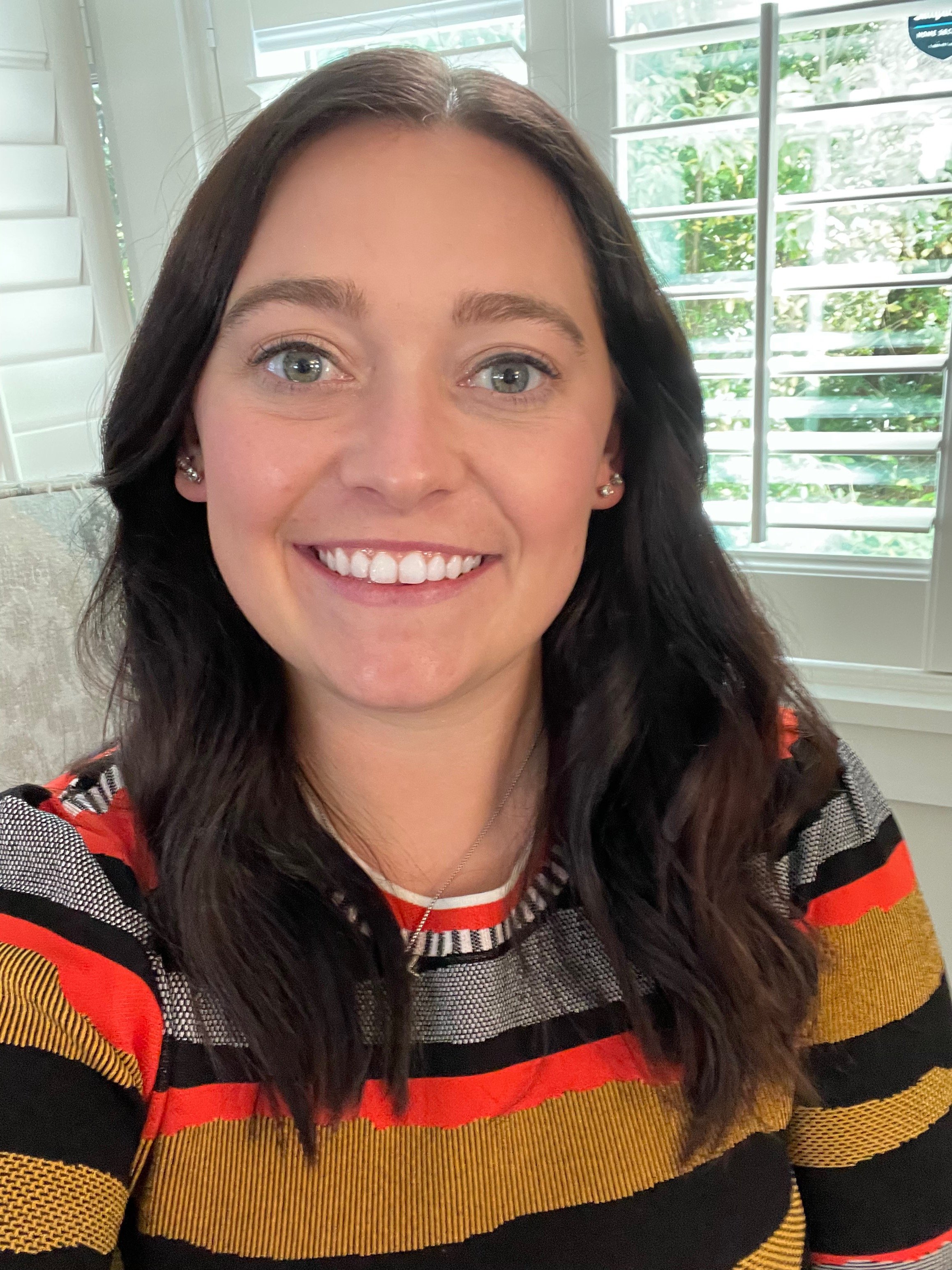 A woman with long dark hair, smiling, sitting in front of a window with blinds and green foliage outside, wearing a colorful striped sweater.