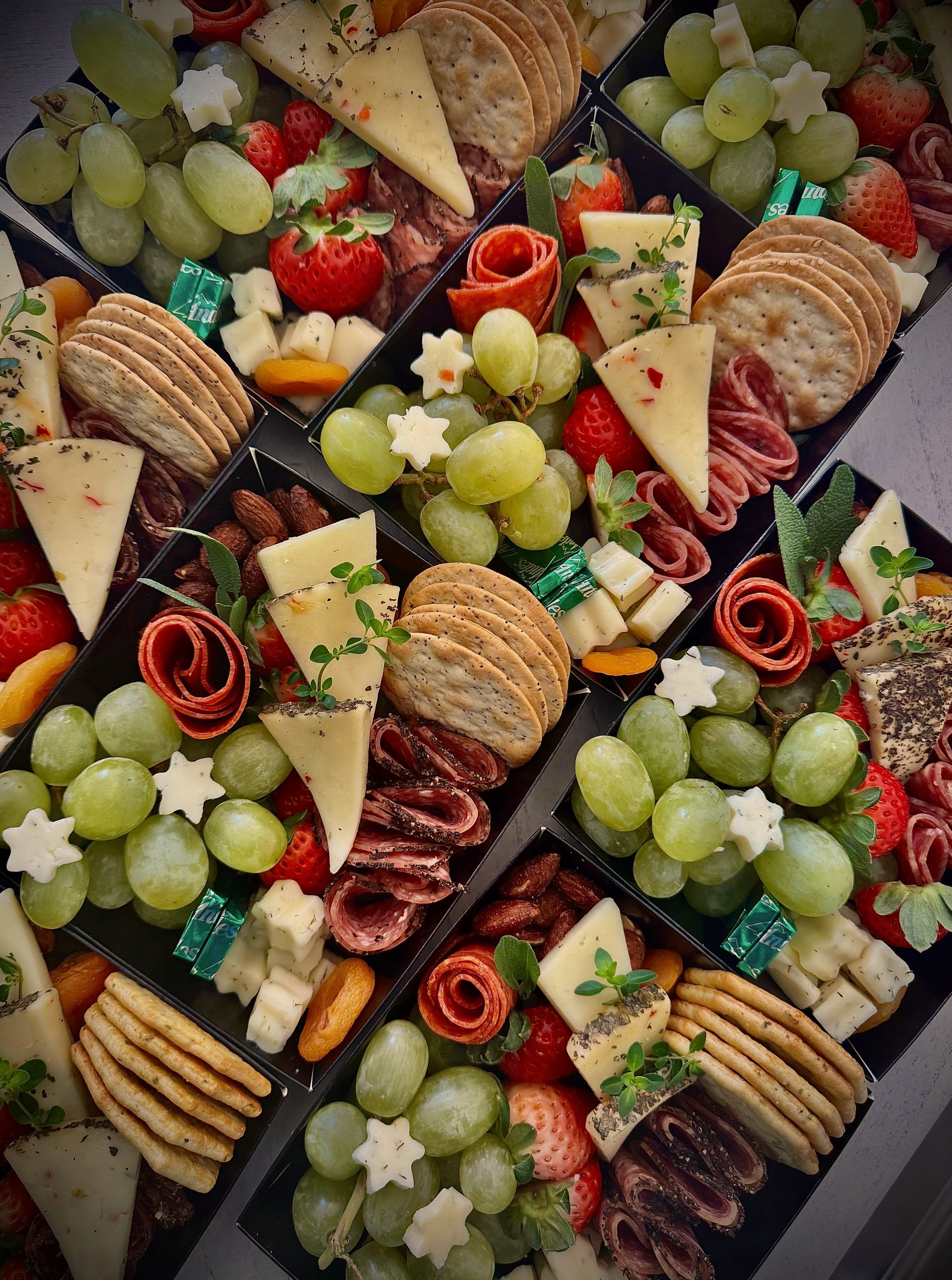 Containers of cut-up fruit, including apples, strawberries, and grapes, on a serving tray with a tray of pudding cups in the background.