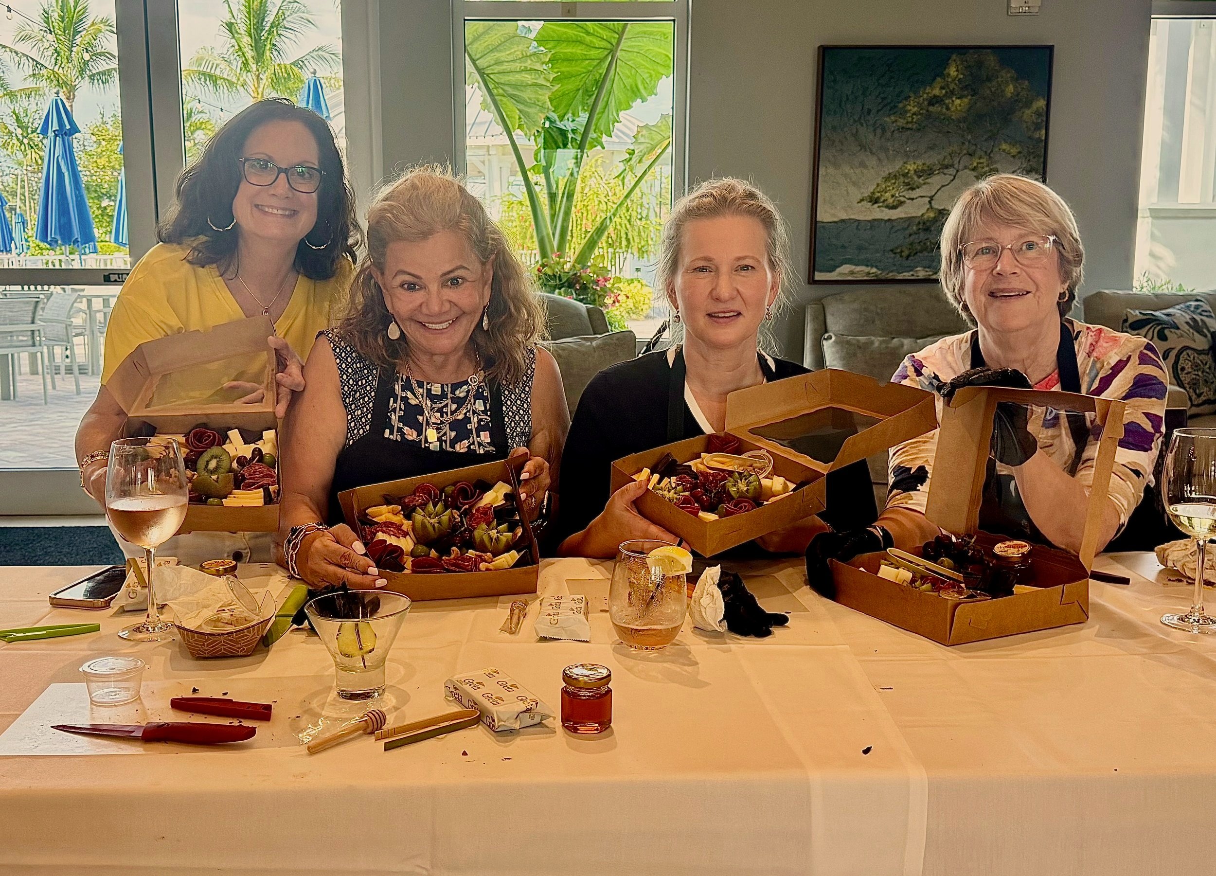 Three women smiling and holding gift boxes filled with flowers, fruits, and chocolates inside a room with a table and chairs, and a window showing greenery outside.