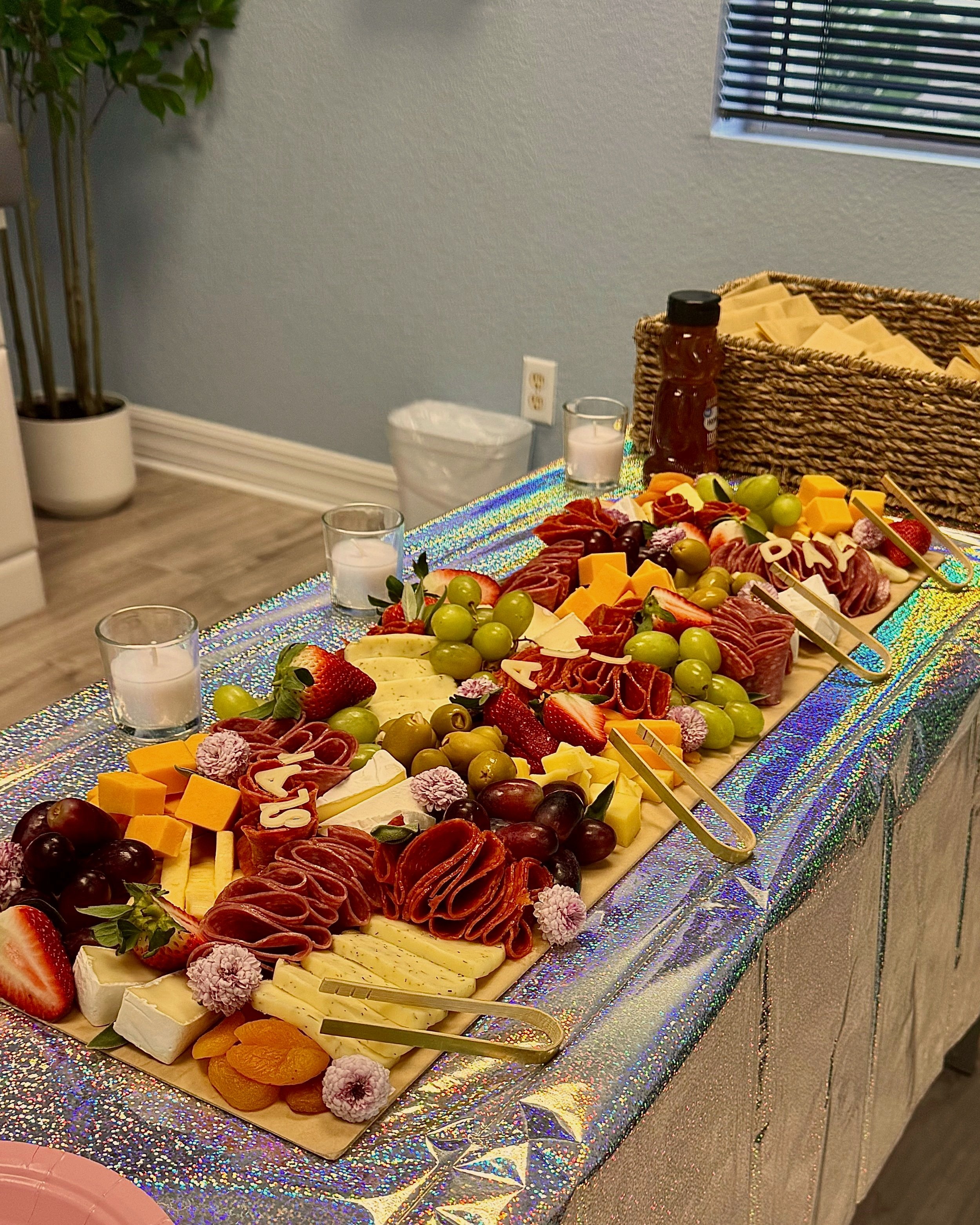 A cheese and charcuterie board with grapes, strawberries, cubed cheeses, sliced meats, and decorative edible flowers on a glittery tablecloth.