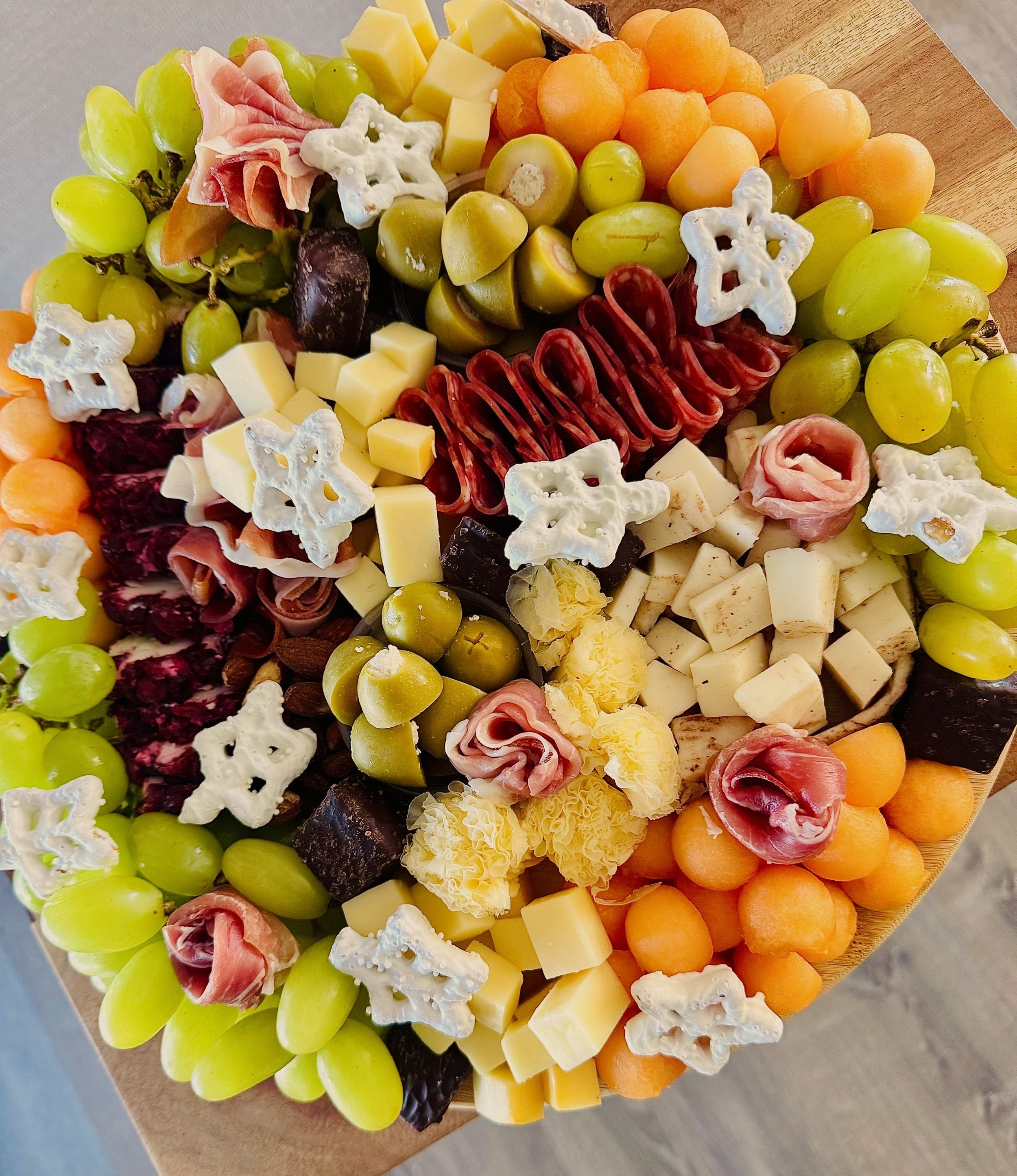 Assorted cheese and fruit platter with green grapes, cantaloupe, honeydew, cheeses, and decorative white cheese crackers on a wooden board.
