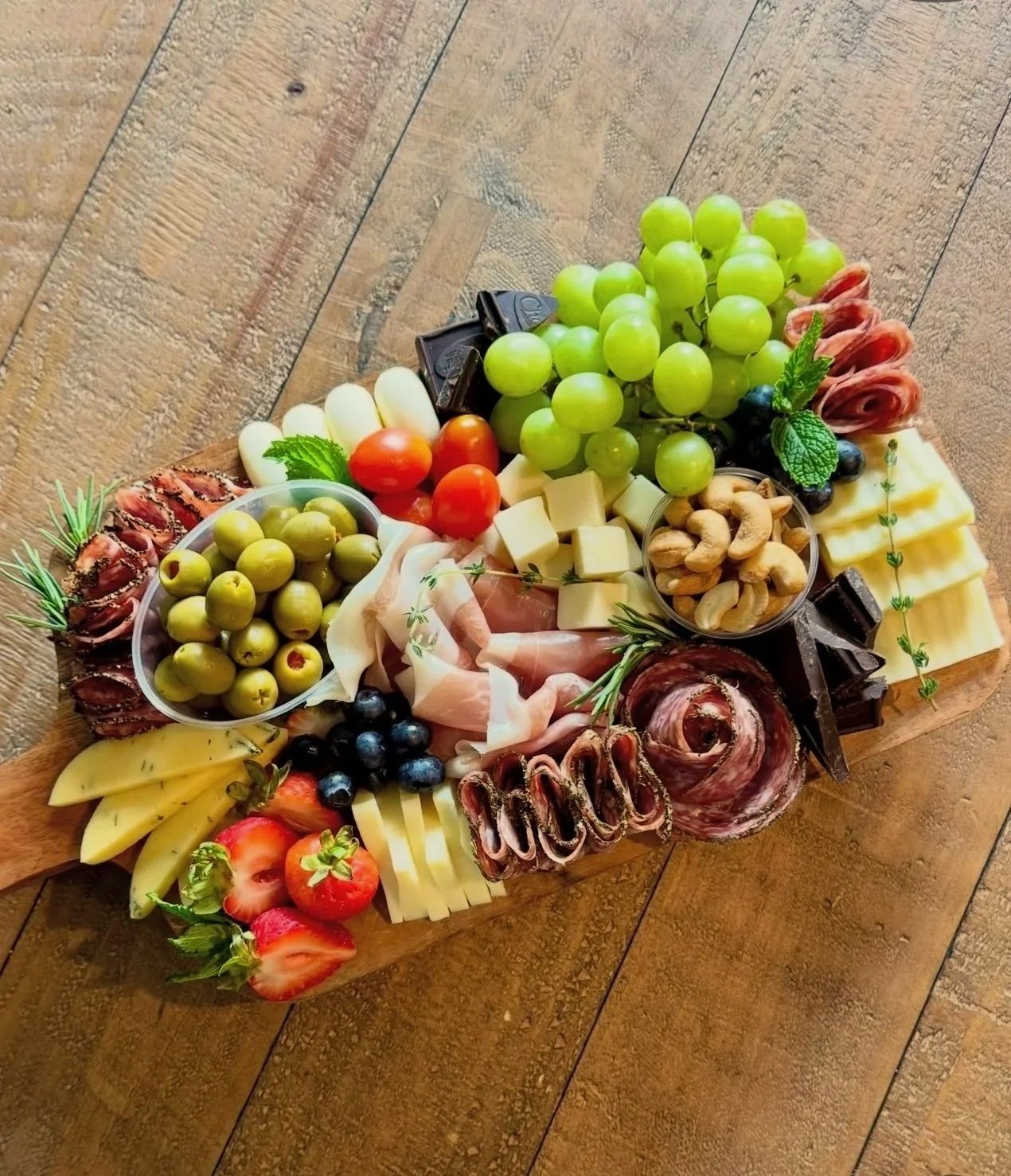 A wooden board with a variety of cheeses, grapes, cherry tomatoes, strawberries, figs, blueberries, green olives, black olives, cashews, prosciutto, salami, and slices of cured meats, garnished with mint, rosemary, and herbs.