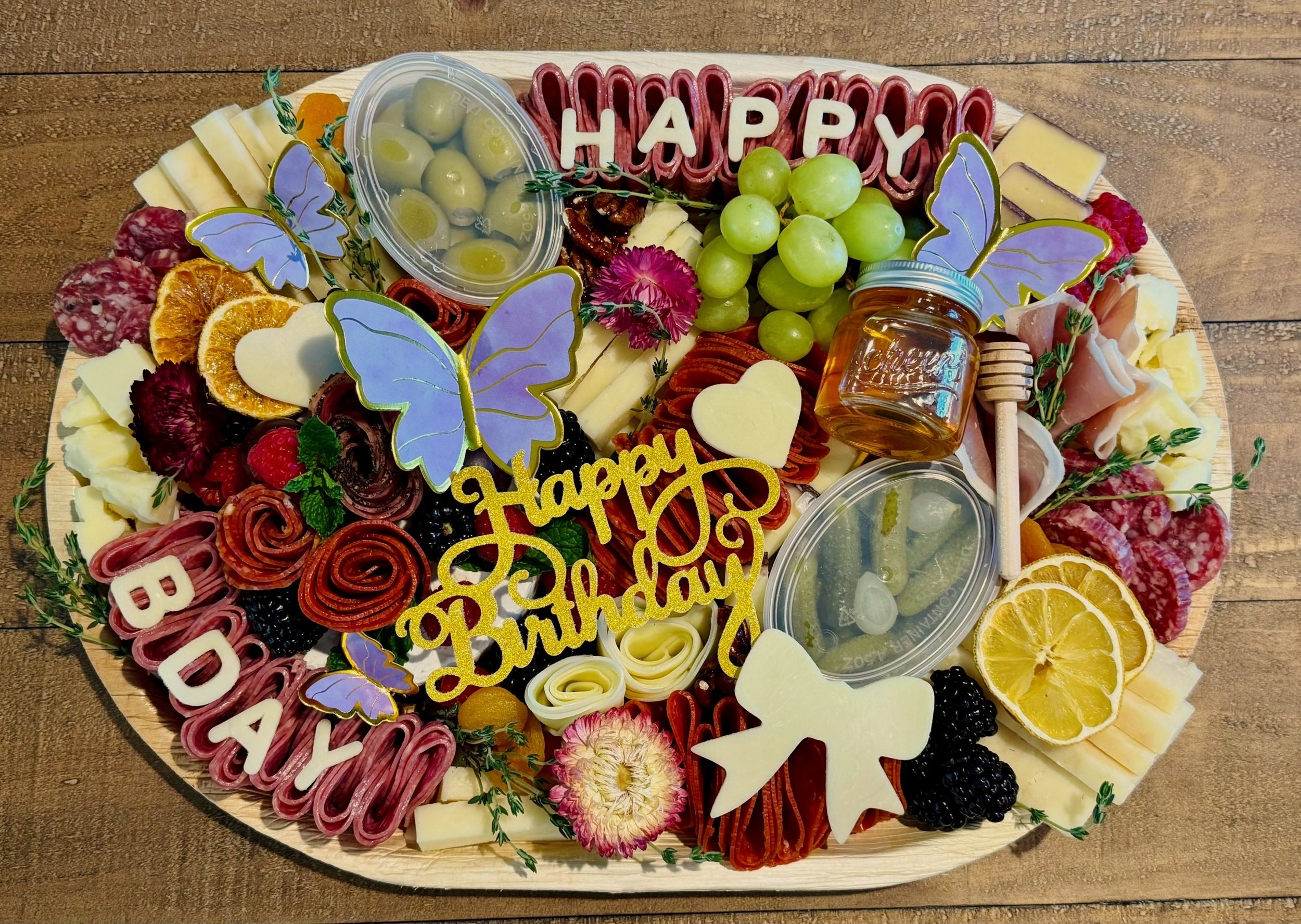 A charcuterie board with green grapes, cherry tomatoes, cheese, crackers, slices of cured meats, green olives, strawberries, and garnishes on a wooden table.
