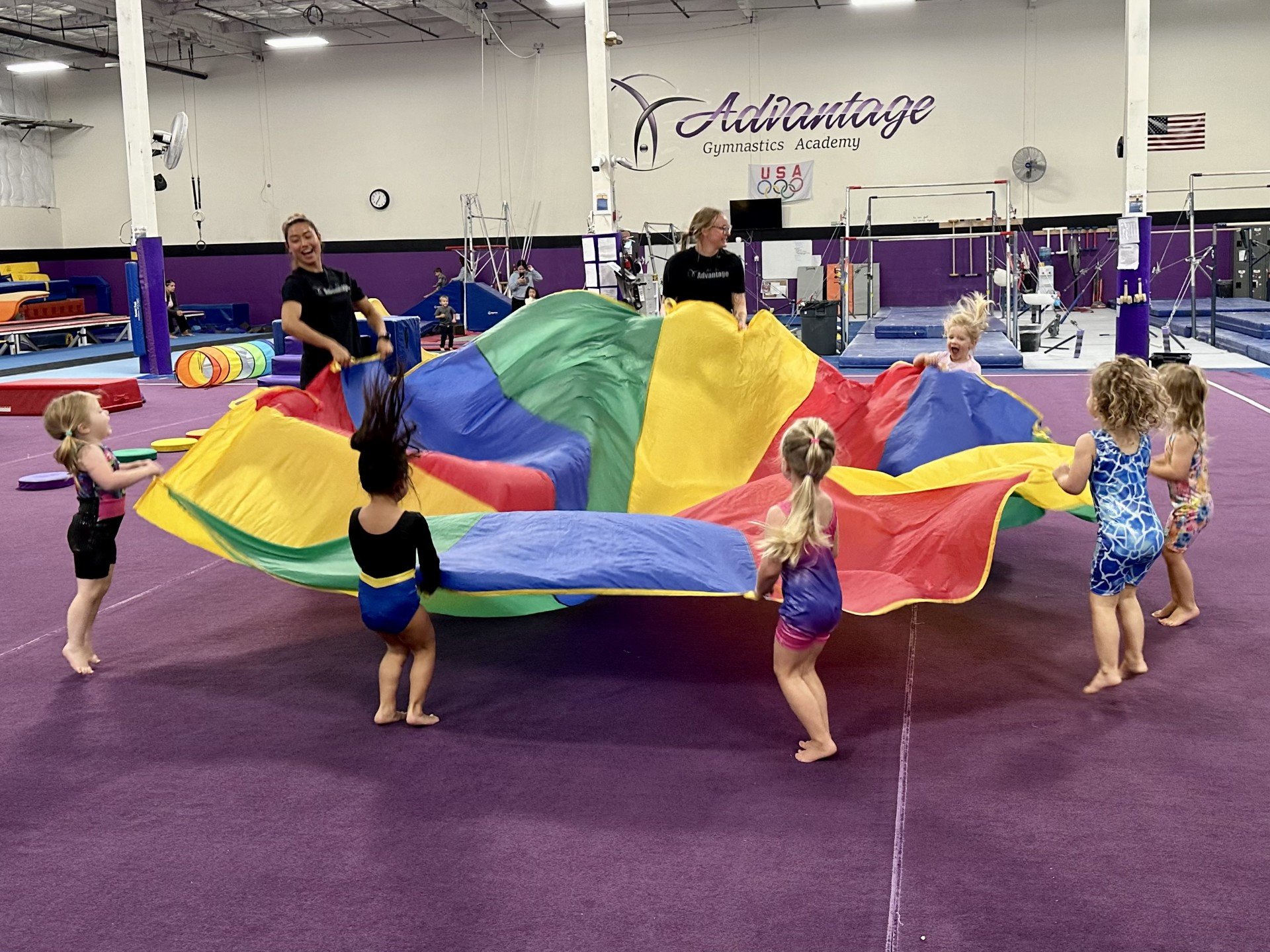Children and adults playing with a large multicolored parachute inside a gymnastics facility.