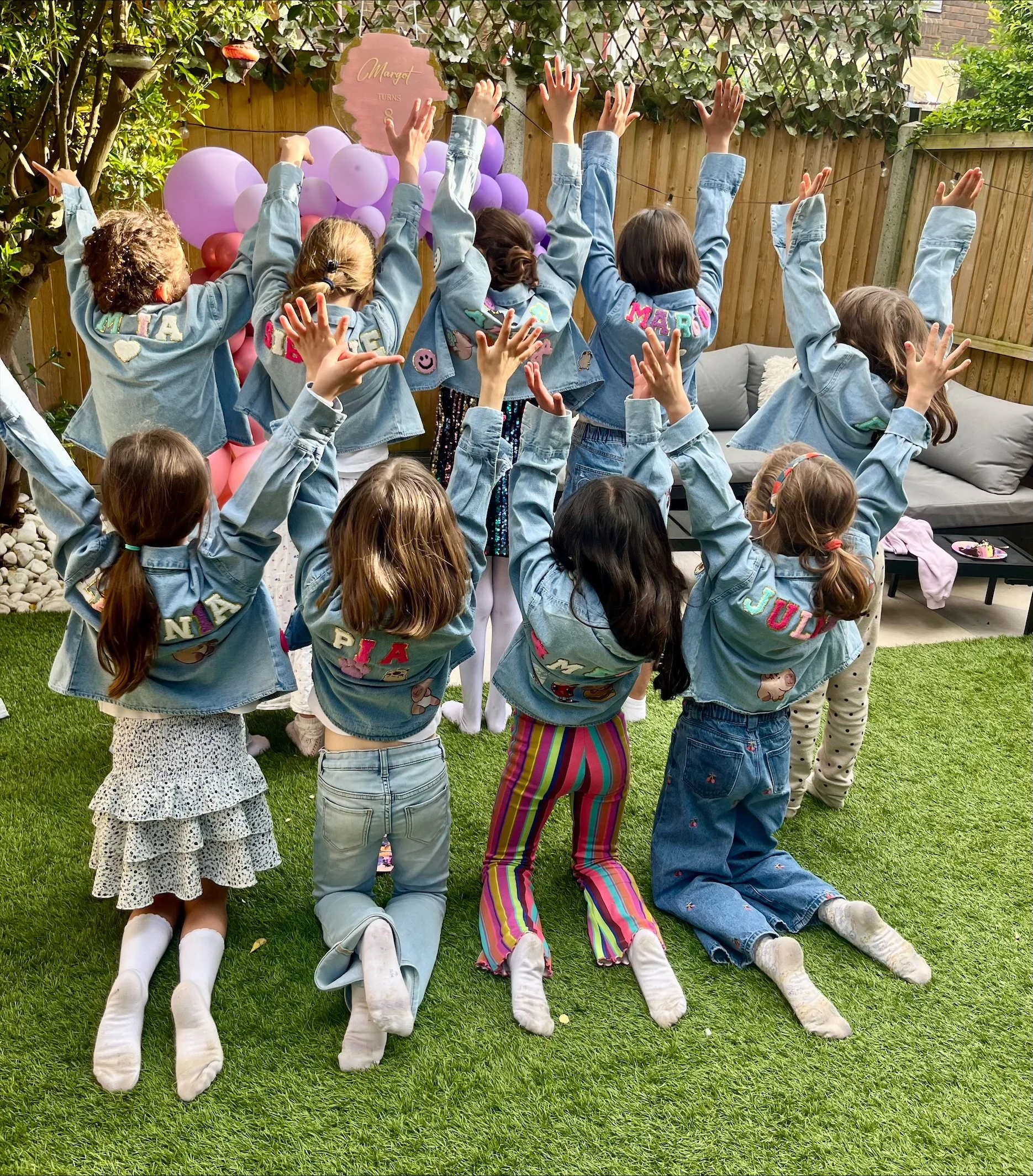 Children lying on grass with legs extended and dressed in colorful clothing, facing away from the camera, during a birthday celebration with pink and purple balloons and decorated signs in the background.