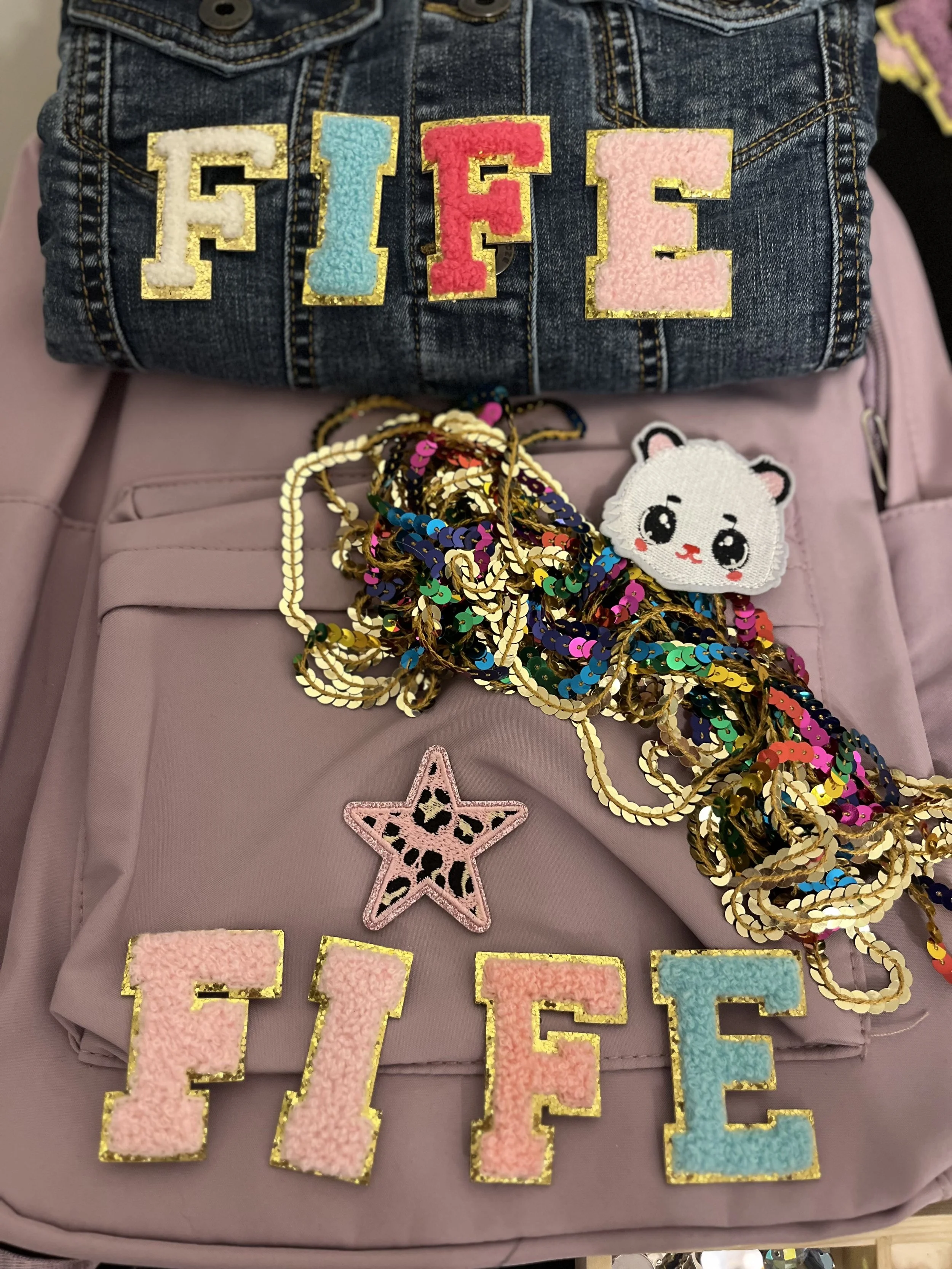Decorative patches of the word 'FIVE' on denim fabric, with various colorful embellishments, small sequin chains, and a cute white embroidered bear face patch with pink cheeks and black eyes.