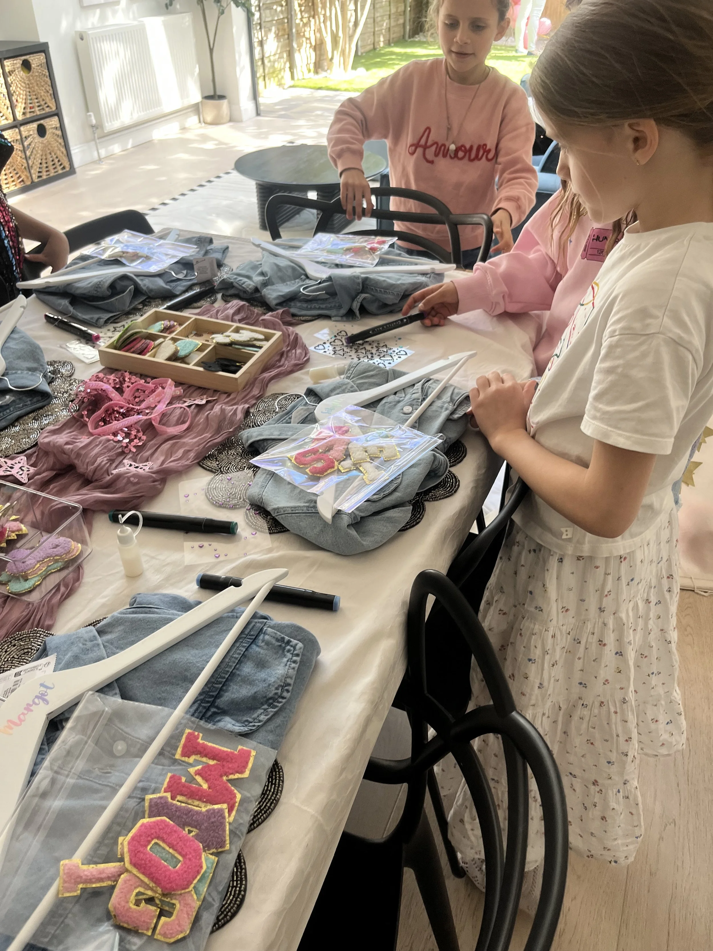 Children gathered around a table filled with fabric, patches, and craft supplies, working on a creative project indoors with sunlight streaming through the window.