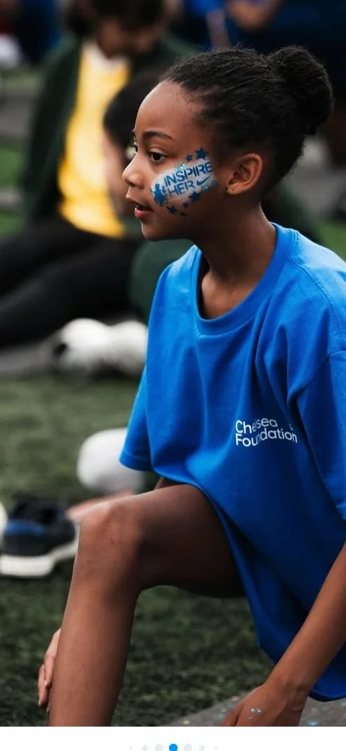 Young girl sitting on grassy field during summer festival, wearing a blue Chelsea Foundation t-shirt, face painted with the words "Inspire Her" and blue stars, with others sitting on the grass in the background.