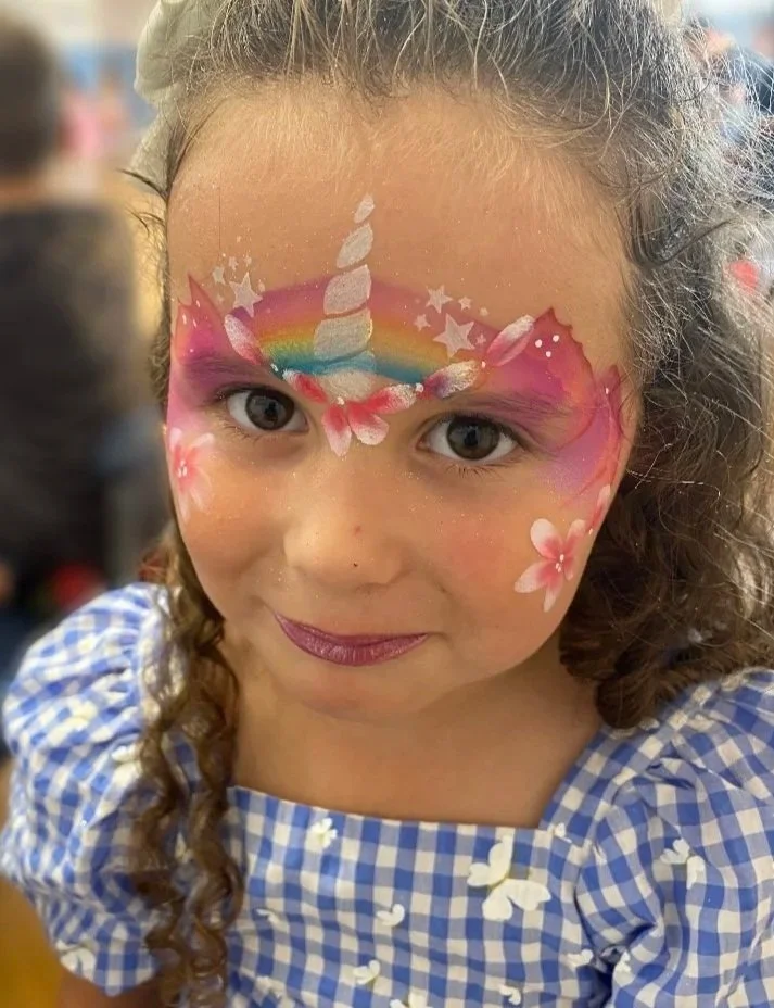 Young girl with curly hair and a blue checkered dress has detailed face paint of a unicorn with a rainbow, stars, and pink flowers on her forehead.