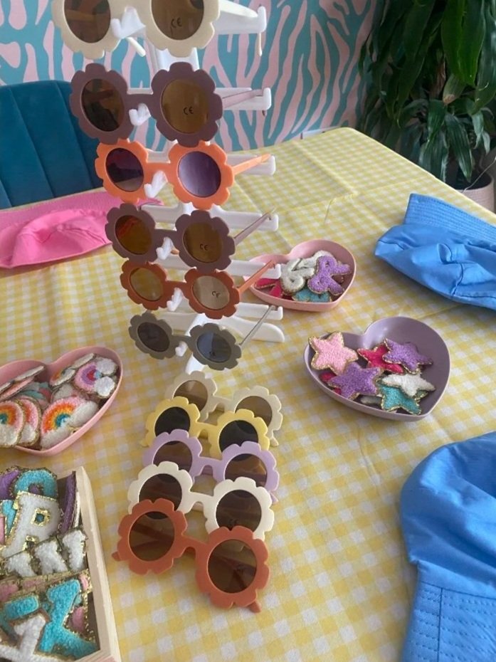Colorful sunglasses arranged on a display stand on a yellow checkered tablecloth, with decorated cookies in the shape of stars and letters, and a pink heart-shaped dish containing more decorated cookies. Blue fabric and a green plant are also visible