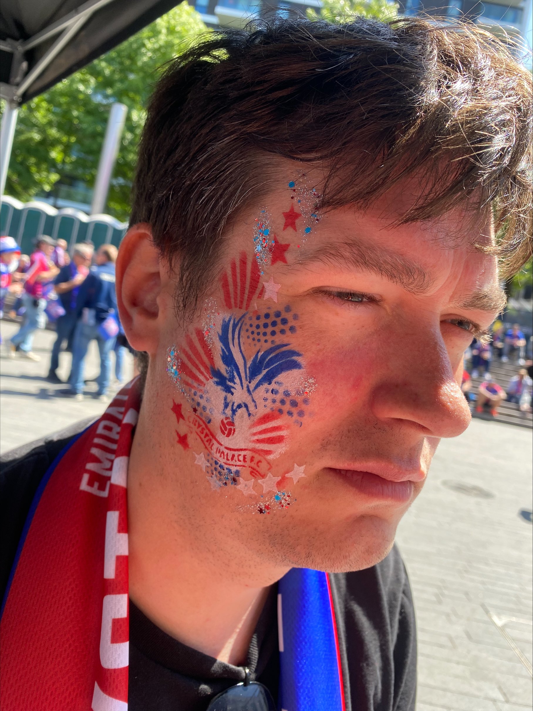 A man with face paint featuring red, white, and blue stars, stripes, and a phoenix with the text 'EUREKA' on his cheek. He is outdoors on a sunny day with a crowd of people in the background.