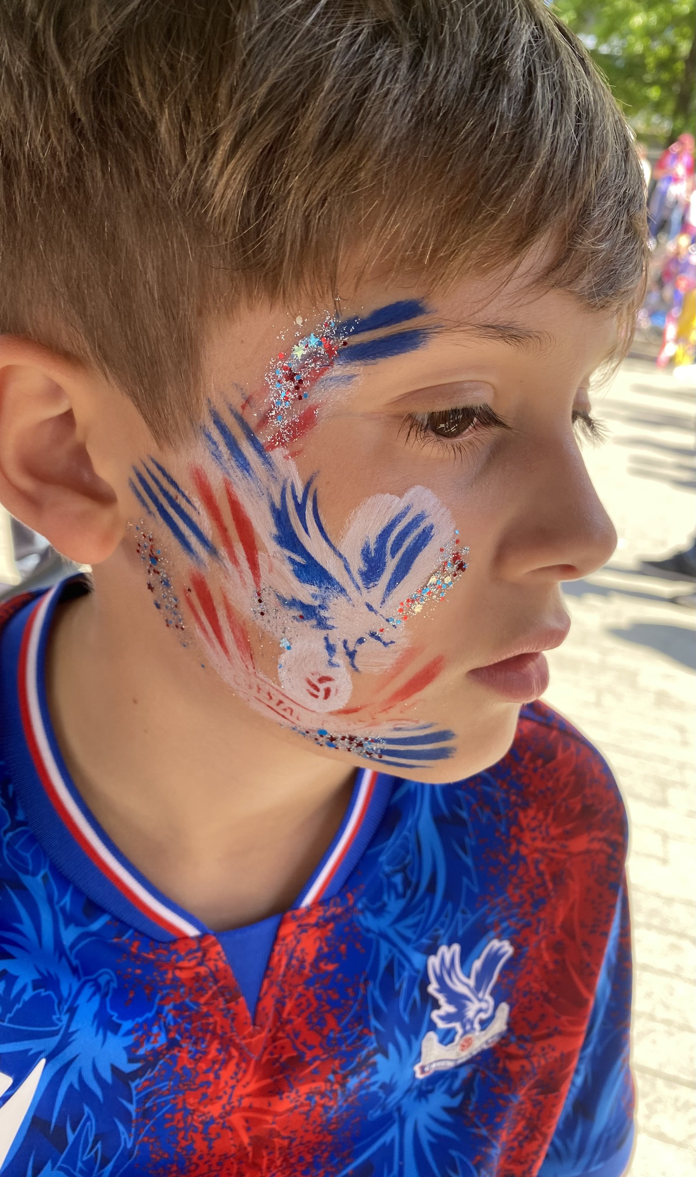 A young boy with face paint in red, white, and blue, depicting a phoenix and the number 5, wearing a blue and red sports jersey.