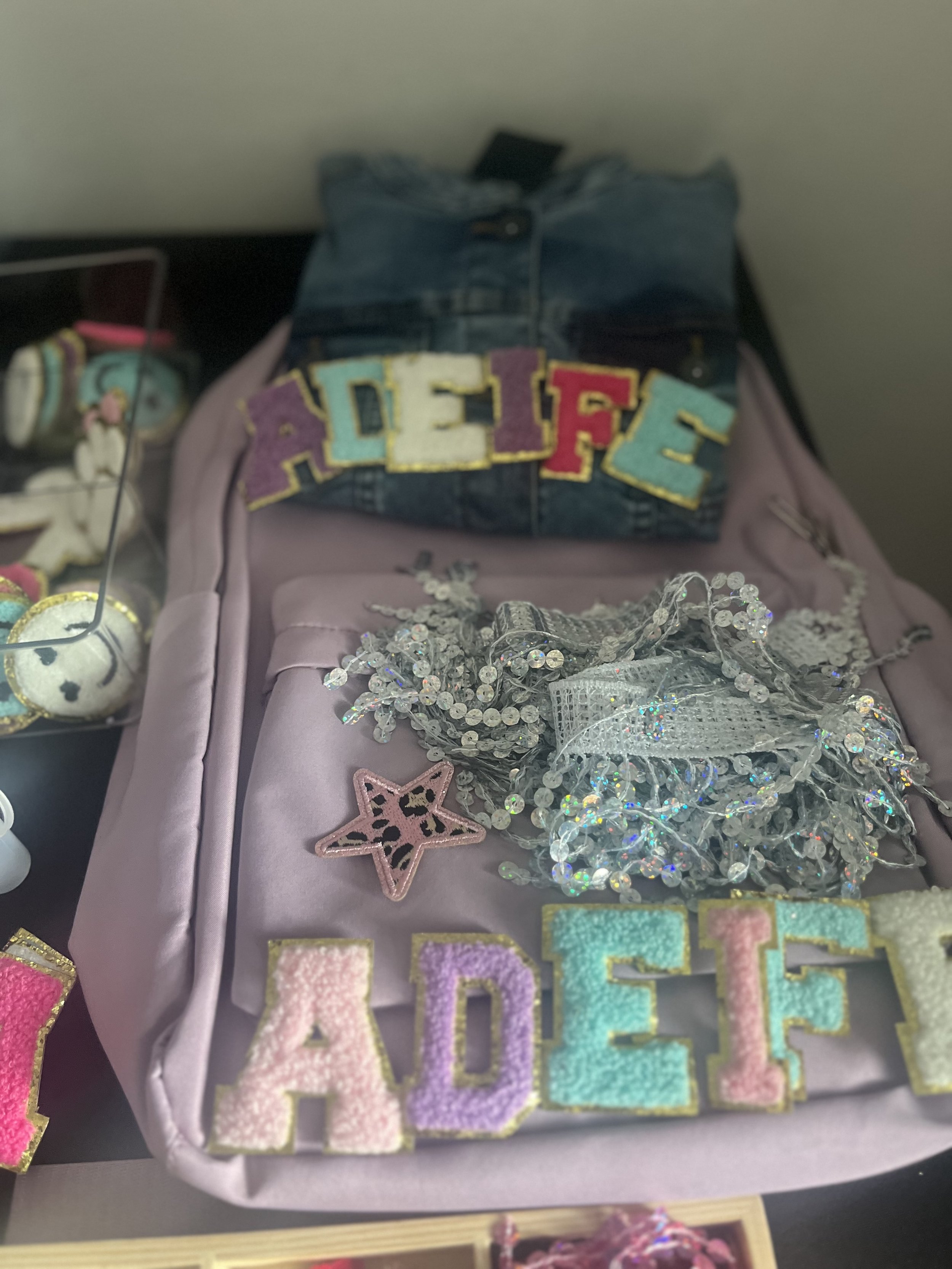 A display of colorful decorative items, including embroidered patches spelling 'BELIEVE,' a pink star-shaped patch with a leopard pattern, a cluster of silver and clear rhinestone jewelry, and various other embroidered patches on a pink fabric background.