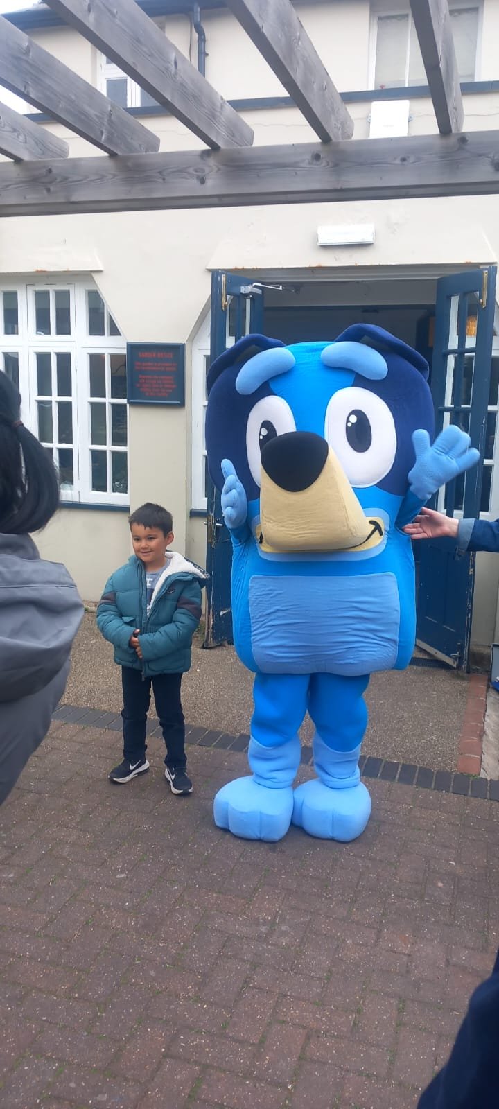 A person in a blue dog mascot costume standing next to a young boy in a blue jacket outside a building with an open door.