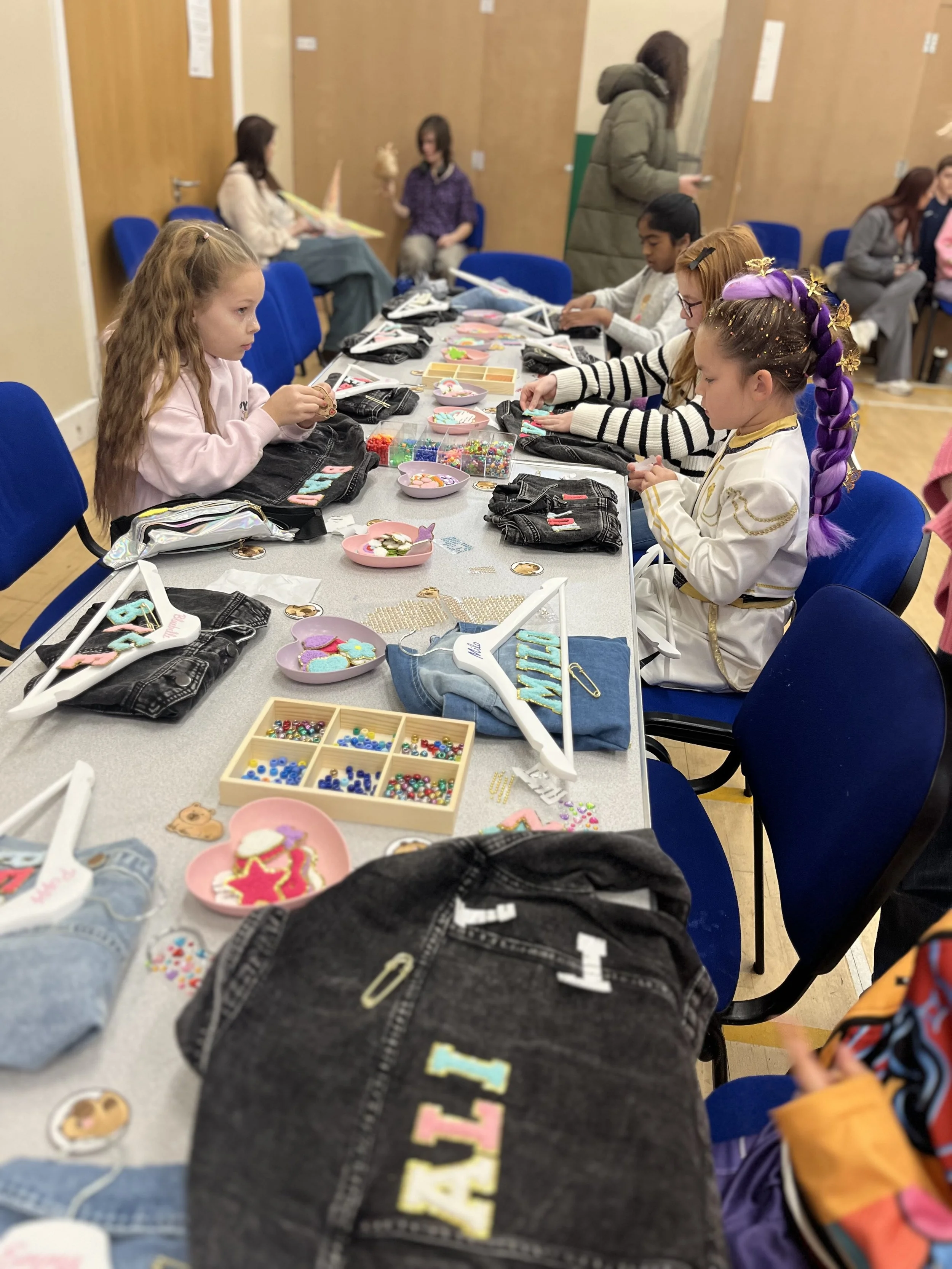 Children participating in a crafts activity, decorating jeans with colorful patches and embellishments in a classroom or activity room.