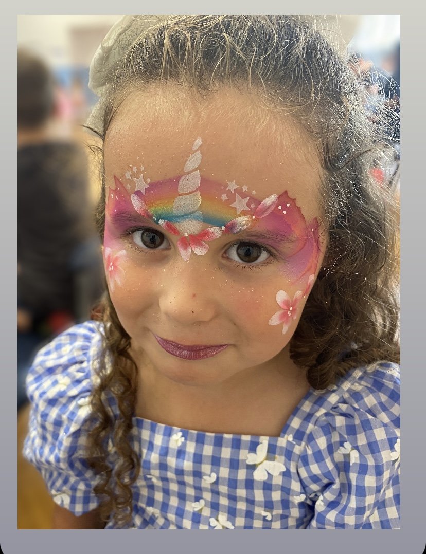 Young girl with curly hair and a blue checkered dress has detailed face paint of a unicorn with a rainbow, stars, and pink flowers on her forehead.