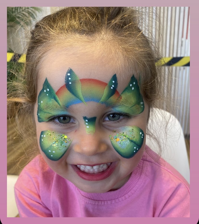 A young girl with face paint resembling a fish with green, blue, and rainbow colors, glitter, and white dots, smiling widely.