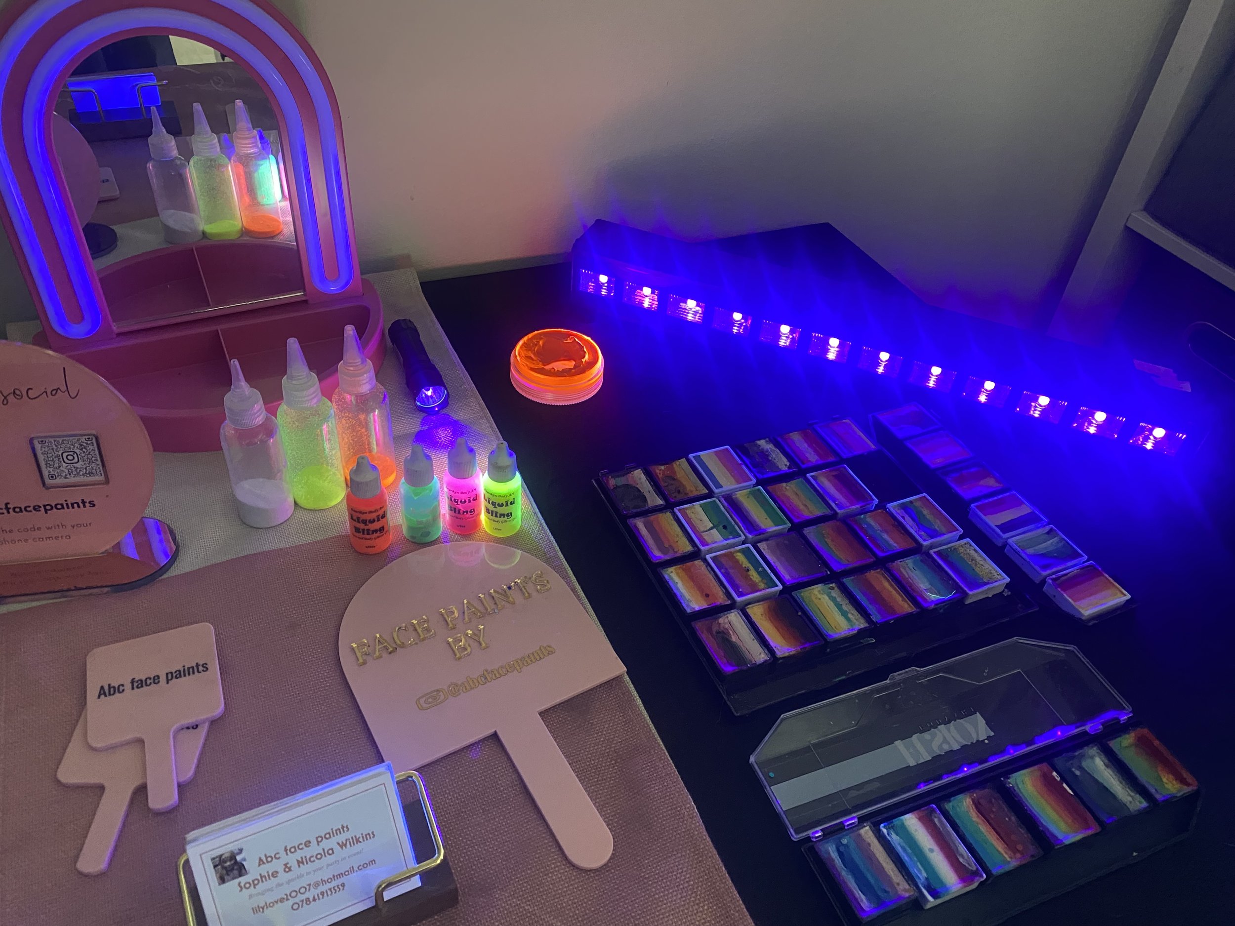 Display of neon-colored face paints and supplies, including bottles, markers, a mirror, and a palette of rainbow face paint colors under purple and blue lighting.