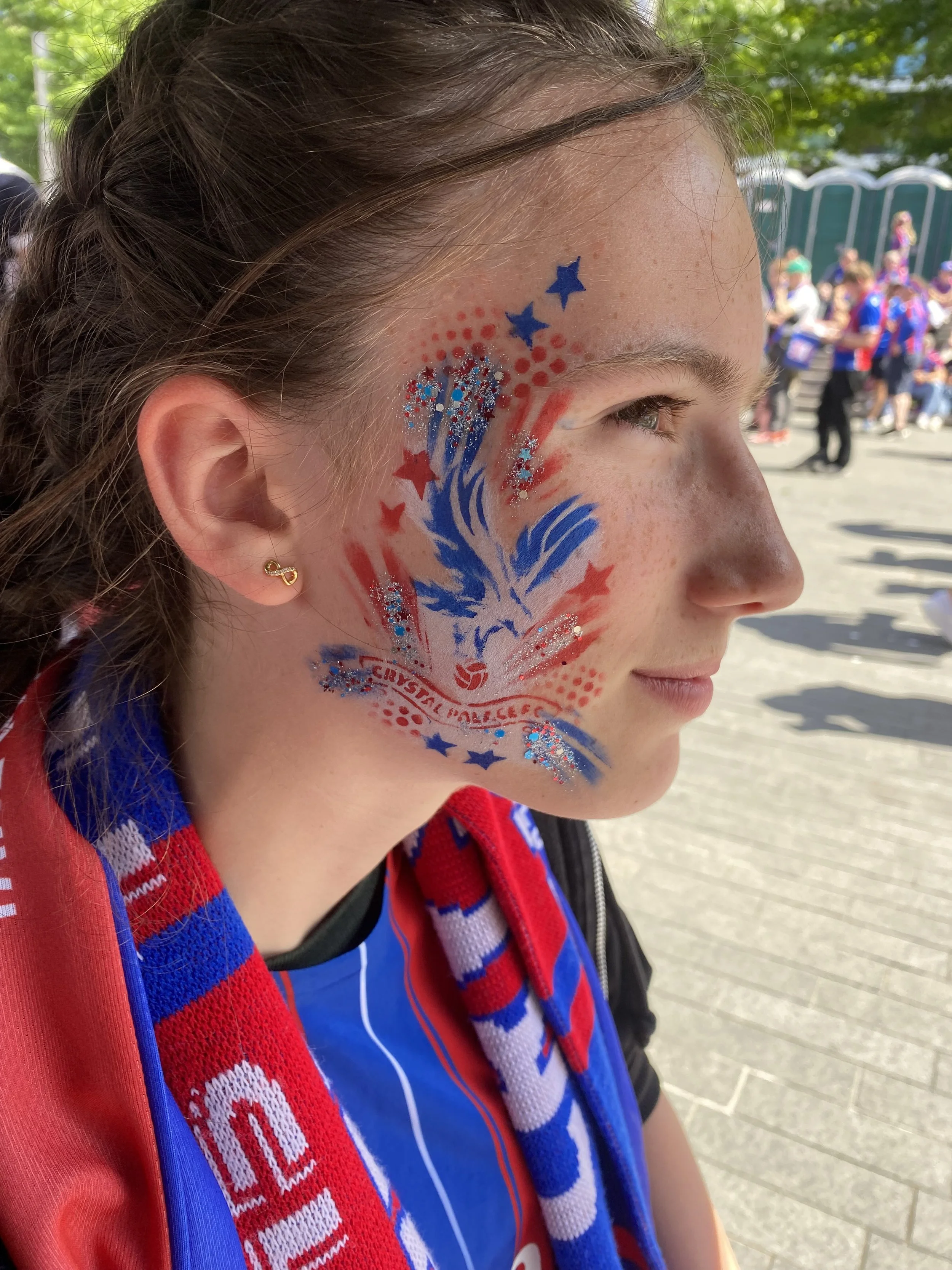 A young woman with a painted face featuring an American flag, stars, and glitter, wearing a red, white, and blue scarf and sports jersey, at an outdoor event.