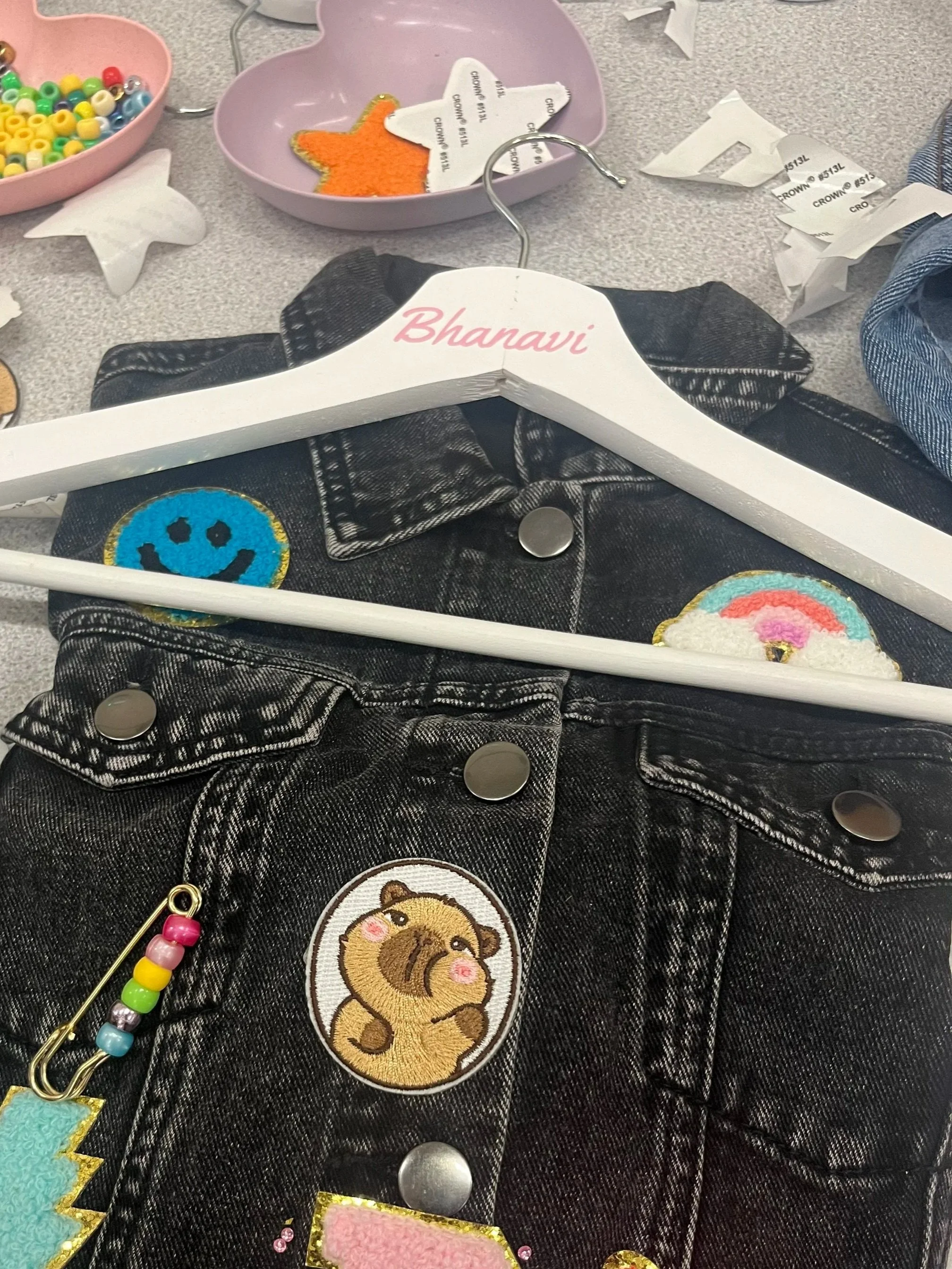 A black denim jacket with embroidered patches, including a brown bear and a yellow lion, on a cluttered grey table. Surrounding the jacket are craft supplies like beads, paper cutouts, and fabric patches.