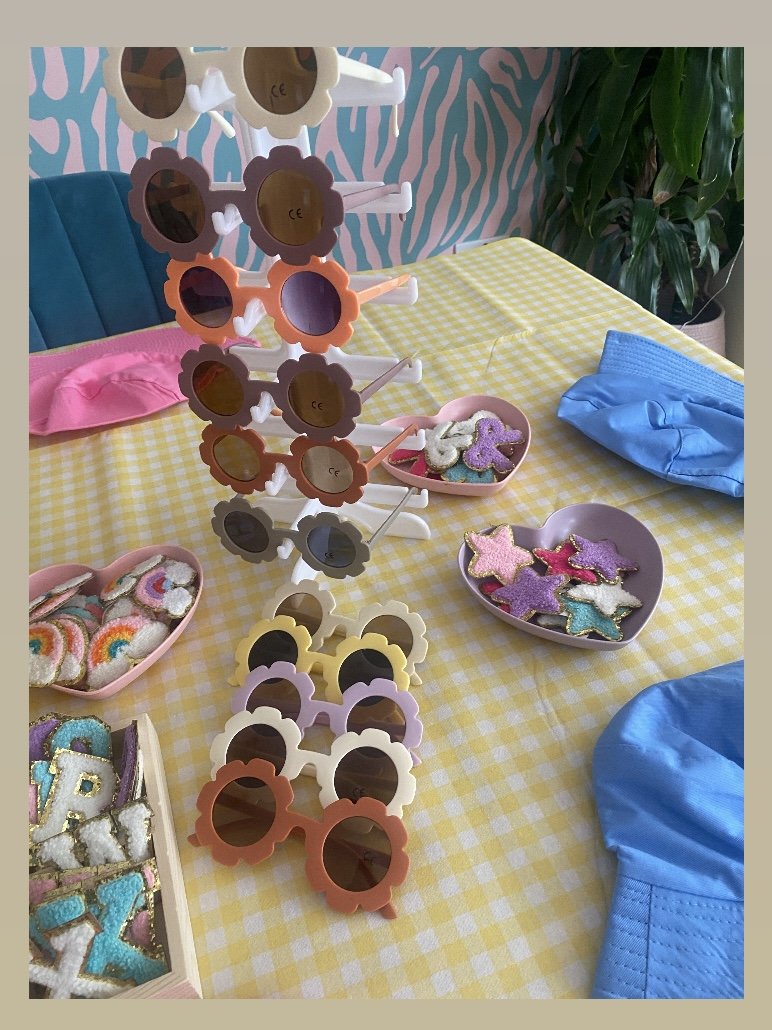 Colorful sunglasses arranged on a display stand on a yellow checkered tablecloth, with decorated cookies in the shape of stars and letters, and a pink heart-shaped dish containing more decorated cookies. Blue fabric and a green plant are also visible