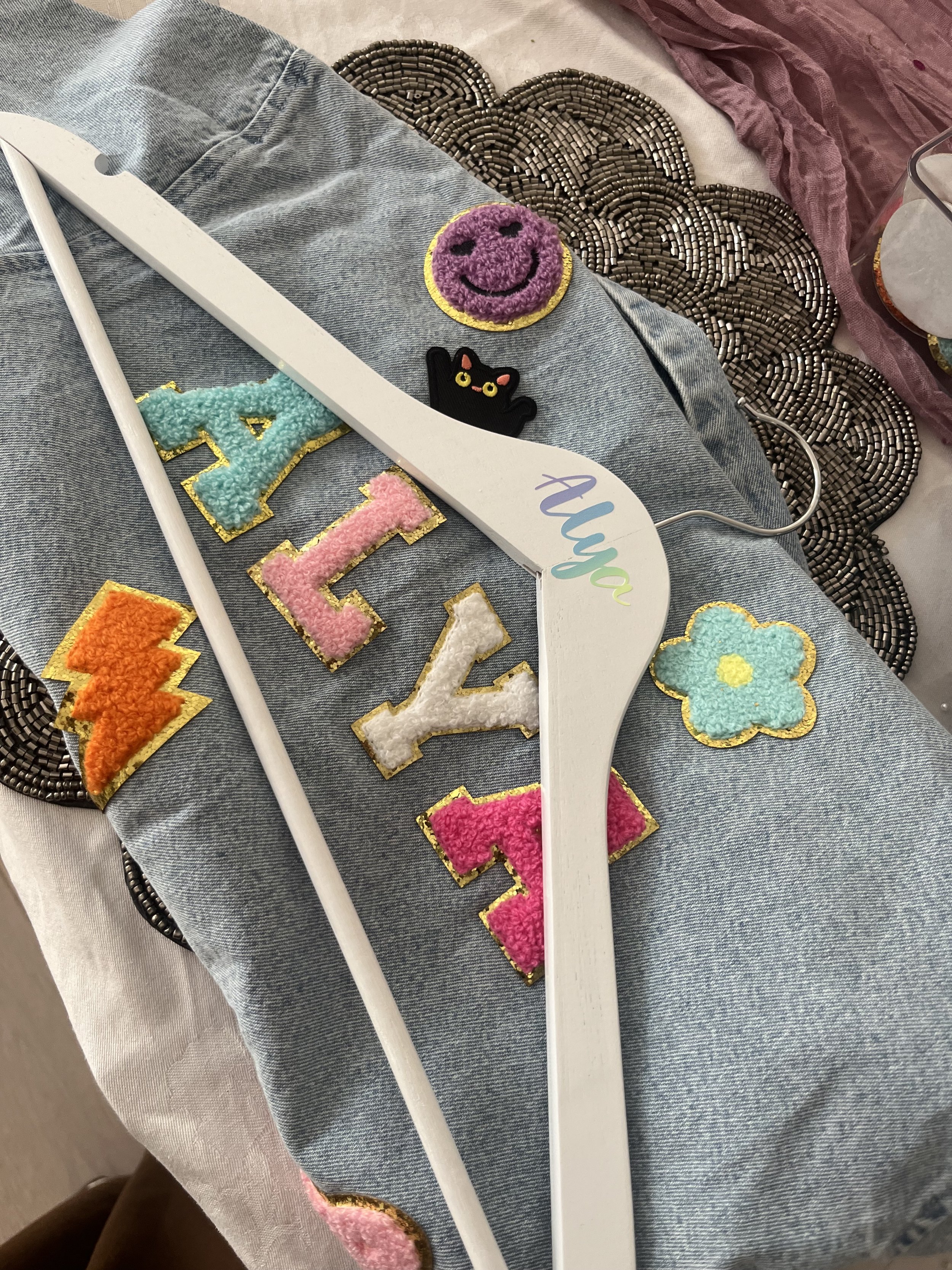 Clothing and craft items on denim fabric, including embroidered patches of a purple smiley face, a black cat, colorful letters, a blue flower, and decorative beaded edges.