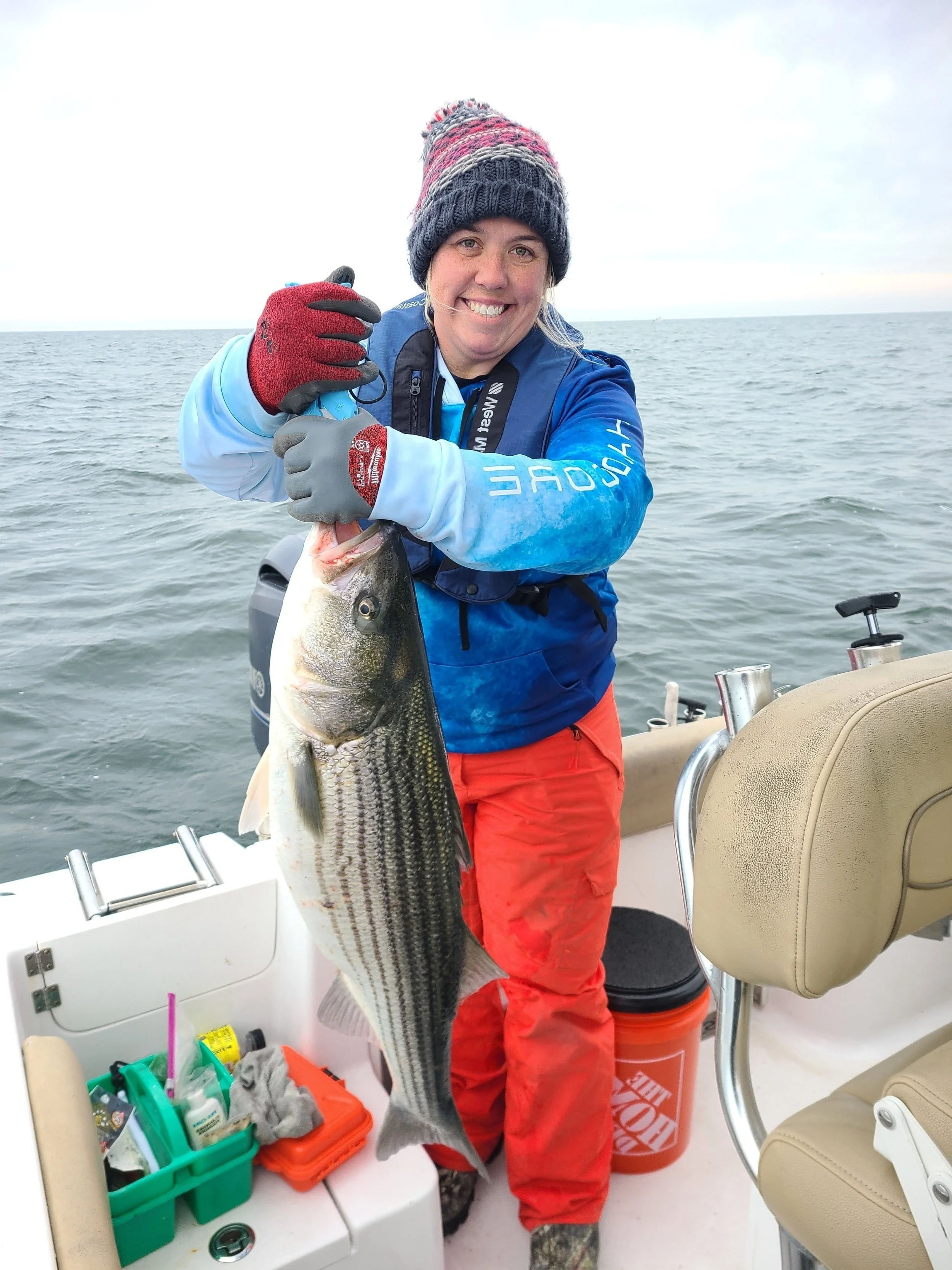 A woman on a boat holding a large striped bass she caught, smiling with water in the background.