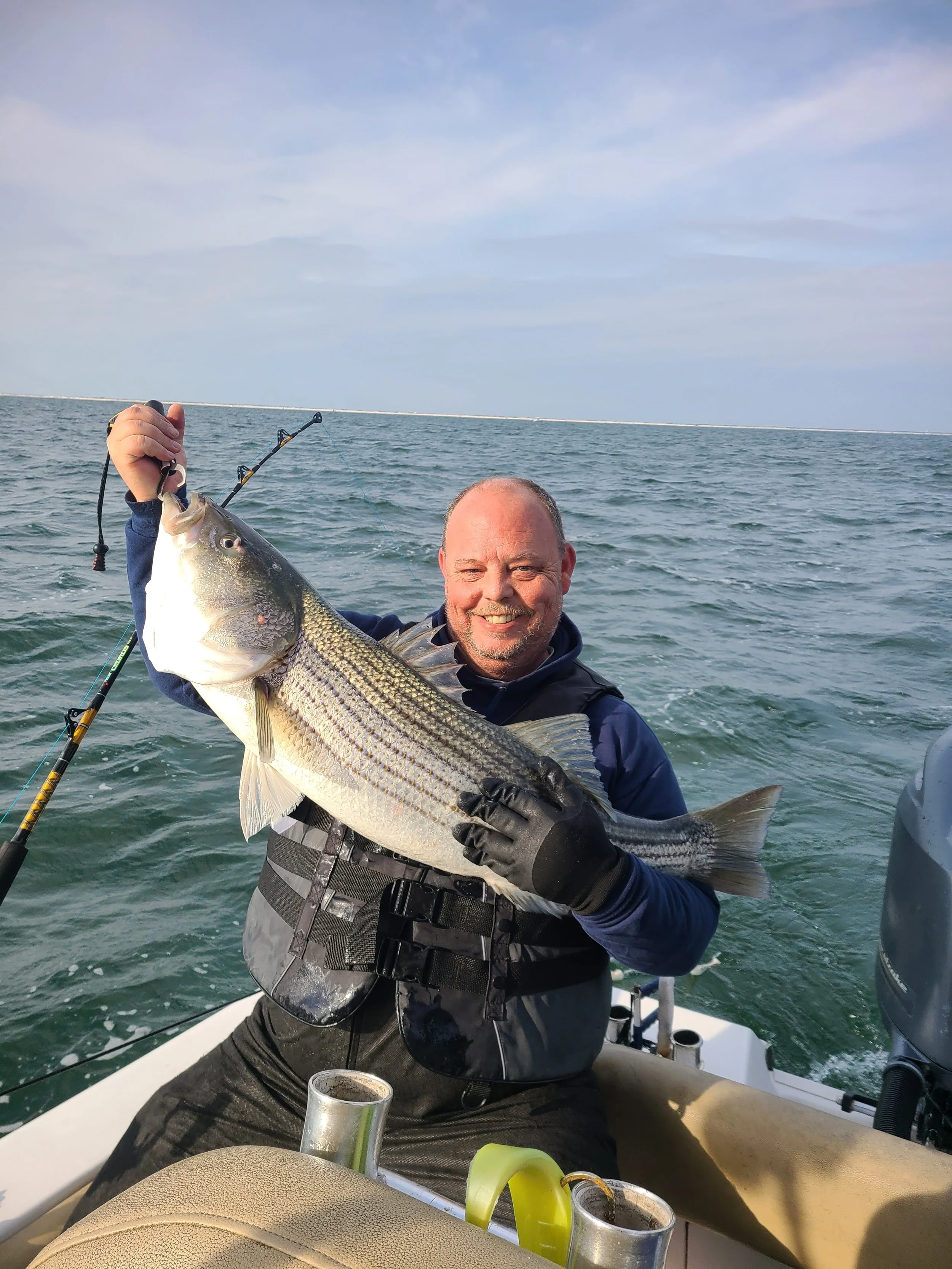 A man on a boat holding a large striped bass he caught while fishing in the ocean.
