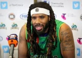 A man with long dreadlocks wearing a white headband and a green sports jersey, speaking into a microphone at a press event.