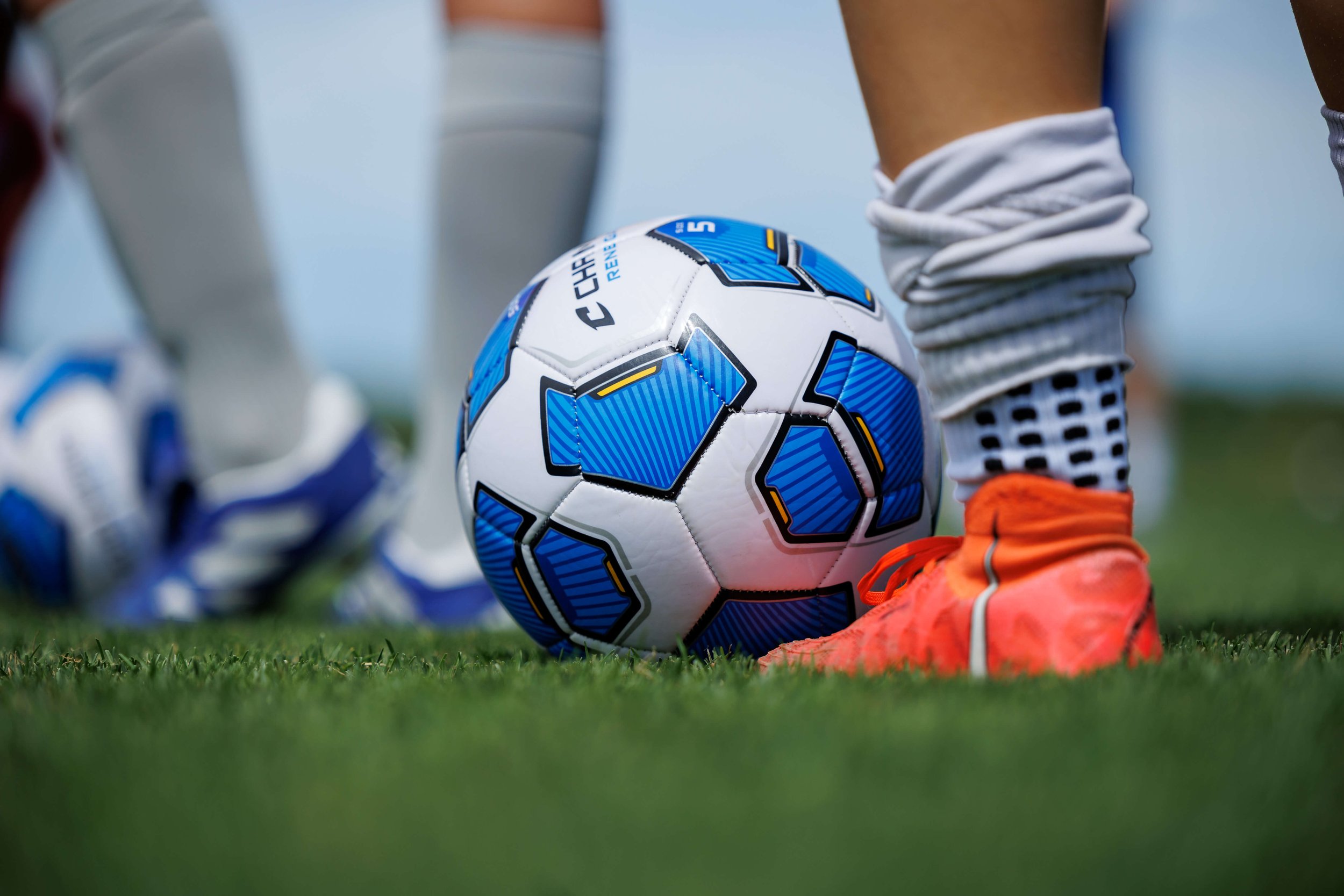 Close-up of a soccer ball on the grass with players standing nearby, one player's foot in bright orange soccer cleats near the ball.