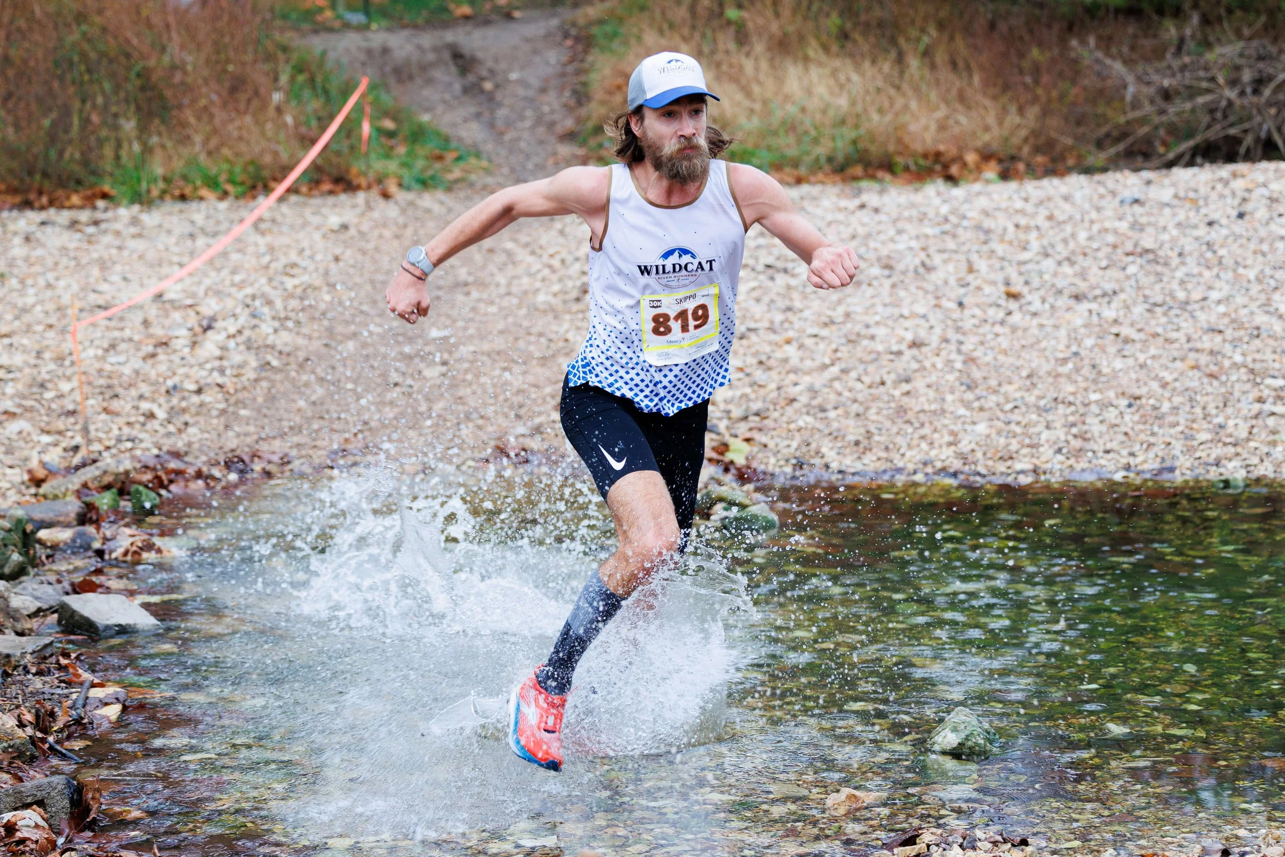 Male runner in athletic gear crossing a shallow creek during a race, with a race bib number 819 and a Wildcat logo on his shirt.