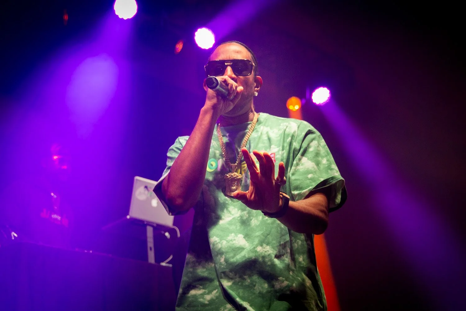 Male performer on stage with purple and orange lighting, wearing sunglasses, a green tie-dye shirt, and gold jewelry, holding a microphone.
