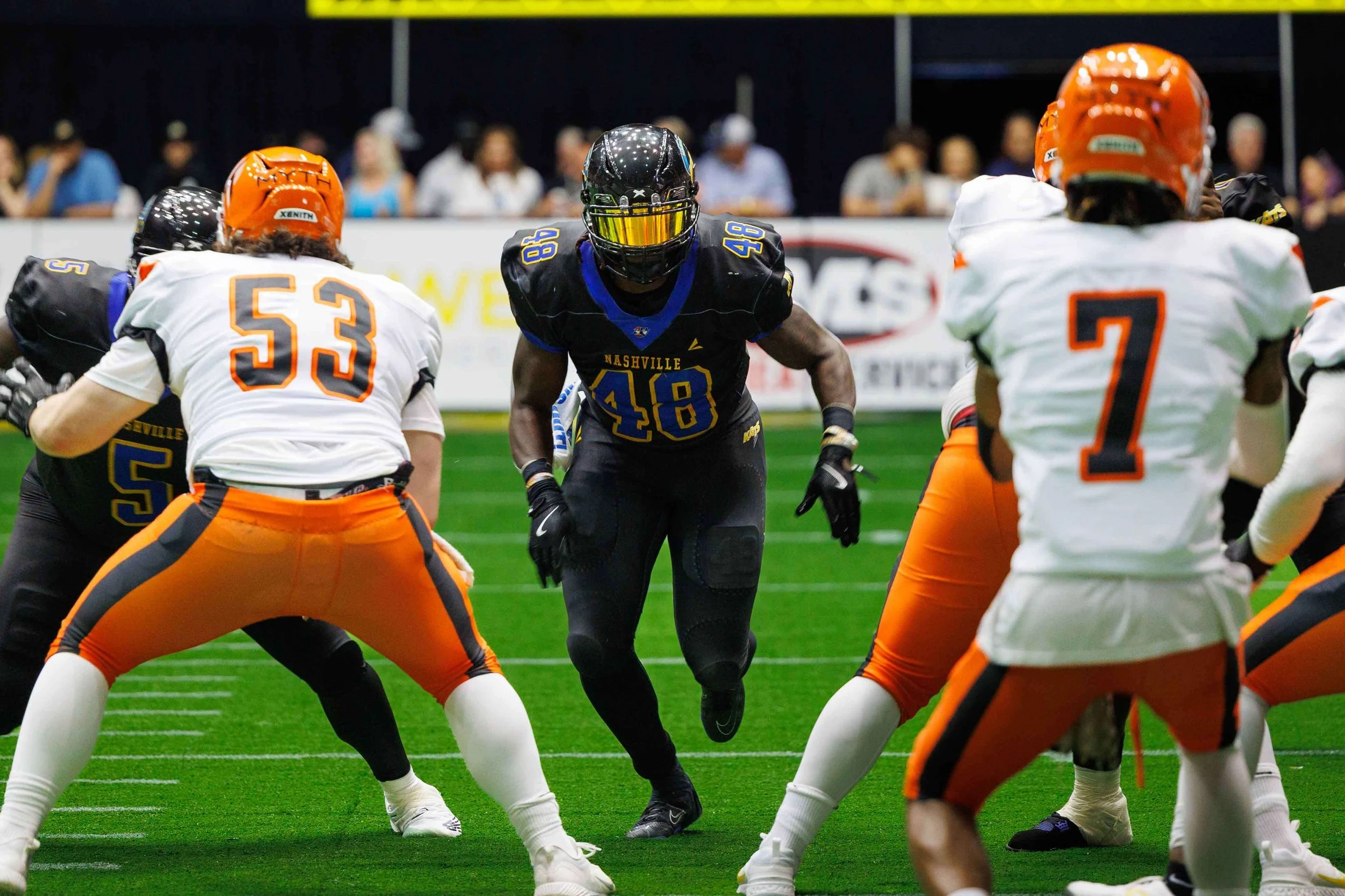 American football players on the field, with one in a black uniform and yellow helmet running, surrounded by players in white and orange uniforms in a stadium.