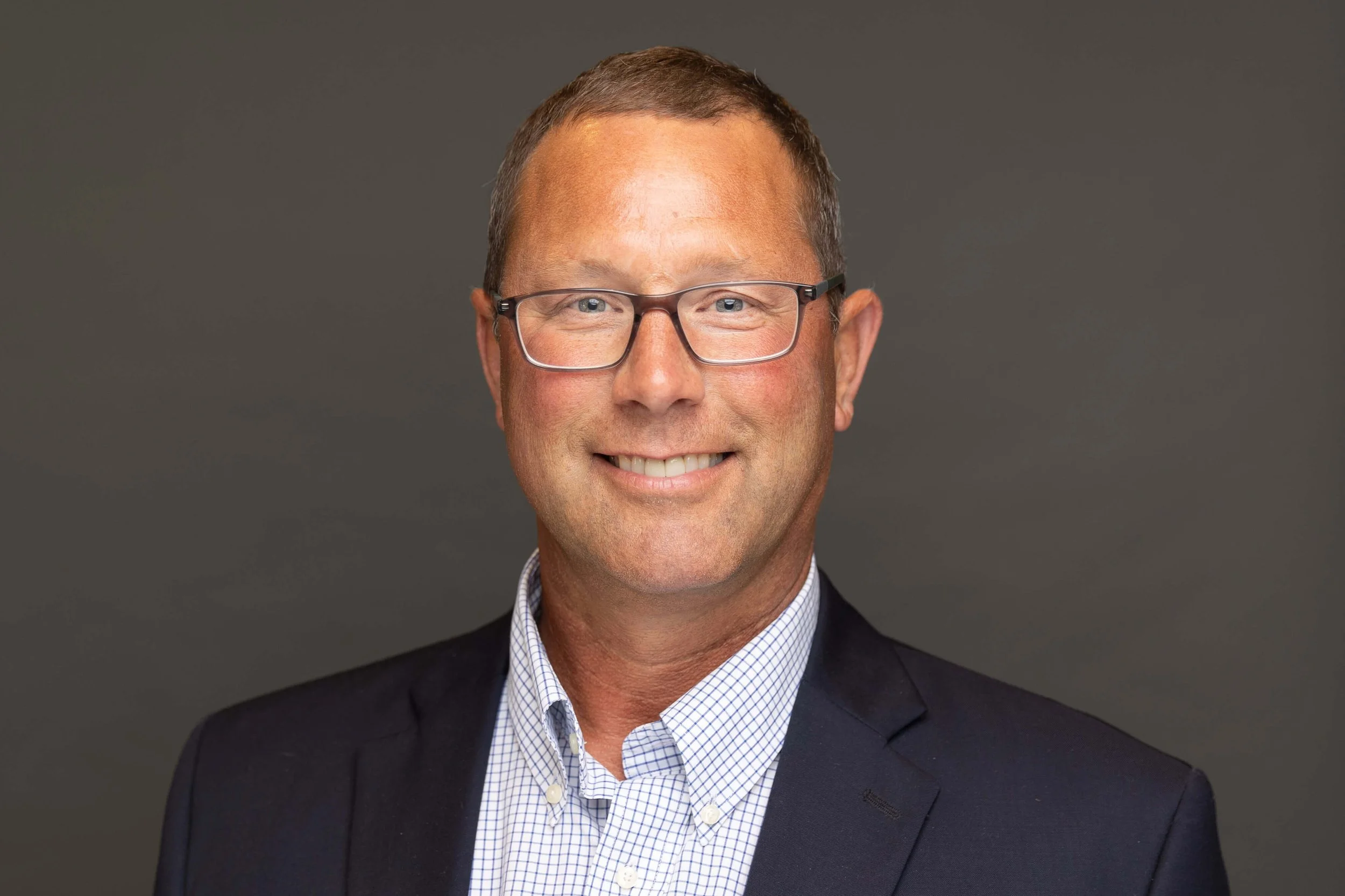 A professional headshot of a smiling man with short brown hair, glasses, wearing a dark suit and a white checkered shirt, against a solid gray background.