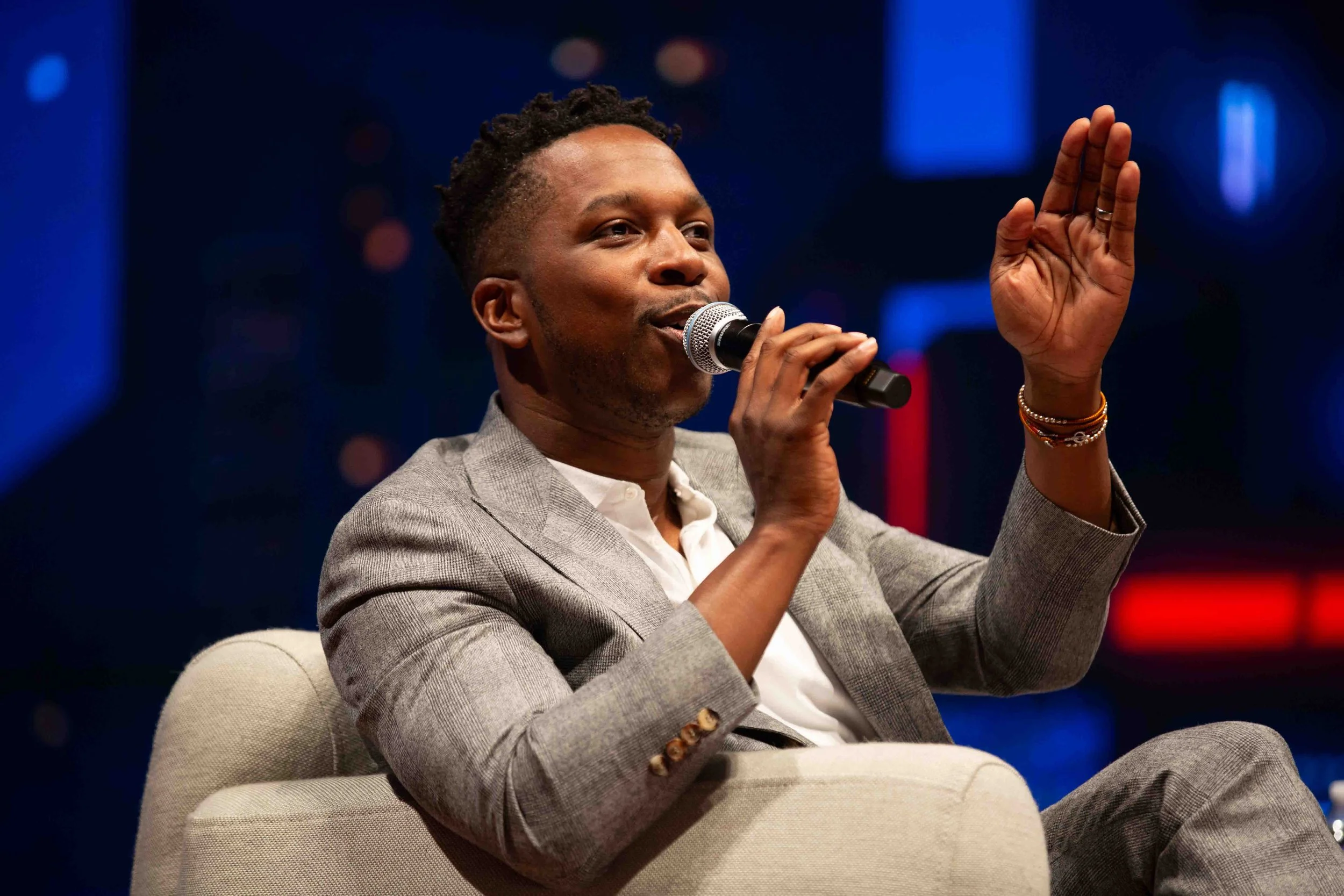 A man in a gray suit holding a microphone, speaking and gesturing with his right hand while seated on a cream-colored chair, against a dark background with colorful blurred lights.