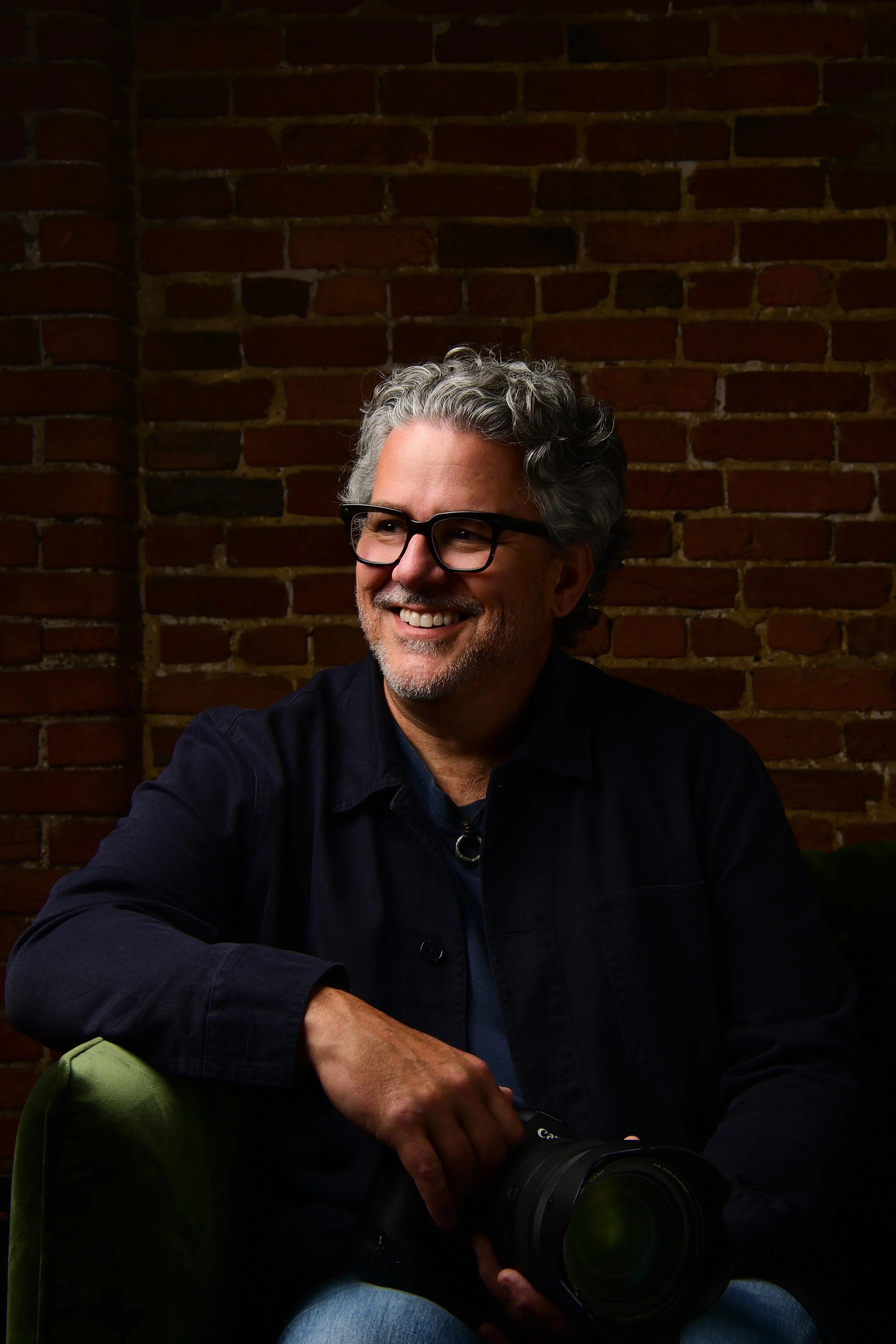 A smiling man with gray hair and beard, wearing black glasses and a dark jacket, sitting on a green chair against a brick wall, holding a camera.