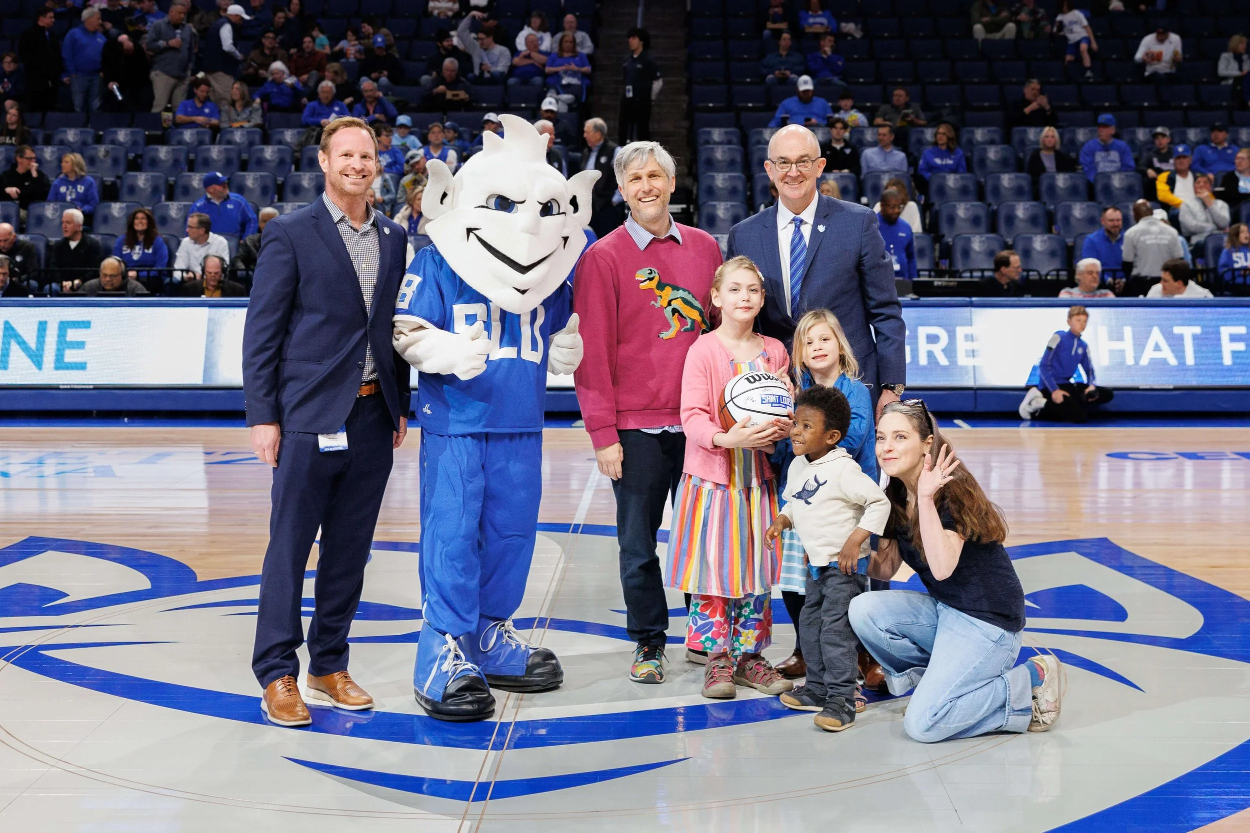 A group of people, including St. Louis University's Billiken mascot, and children, posing for a photo on a basketball court inside a stadium with spectators in the background.