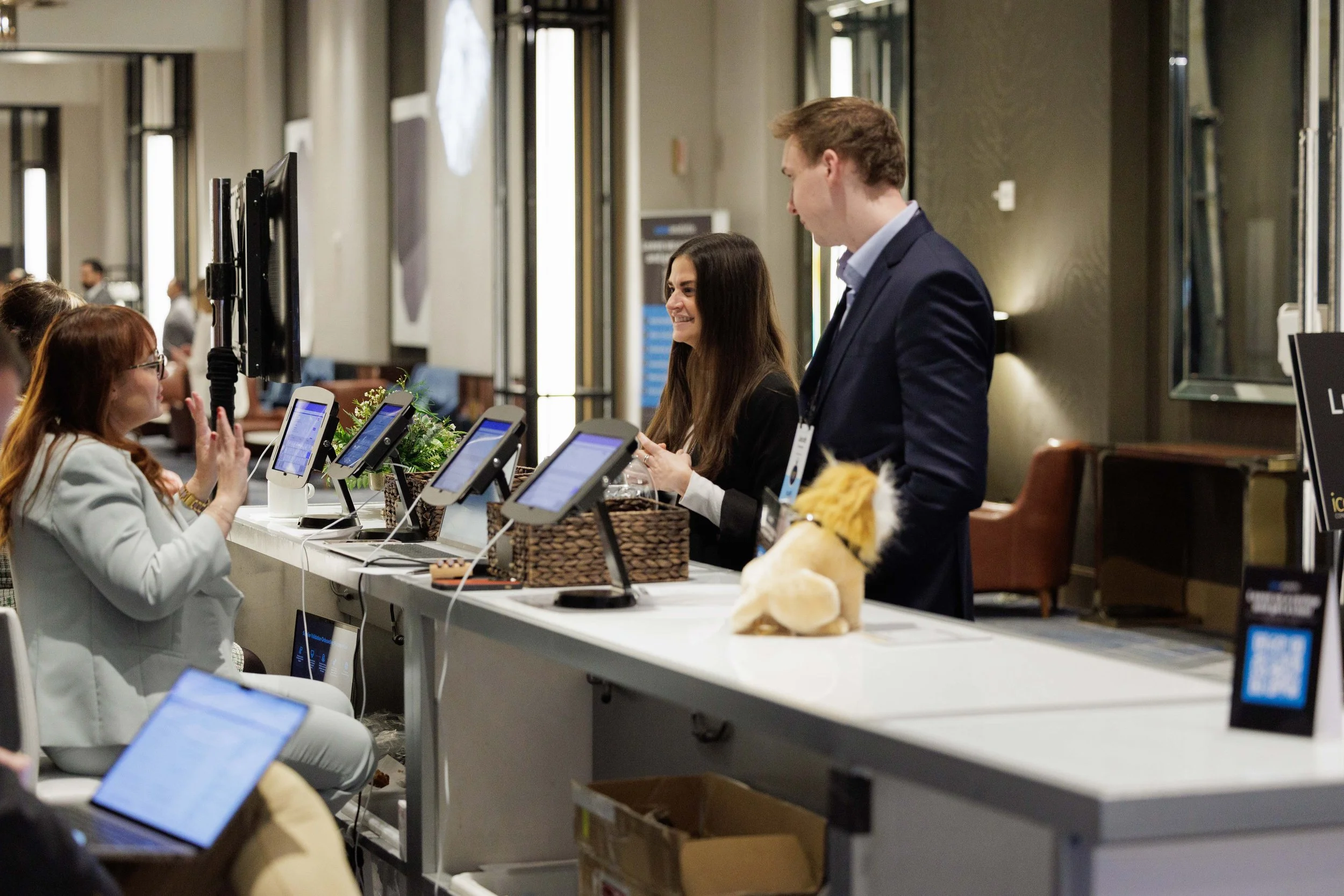A registration desk at a conference or event where a woman with long dark hair and a man with short light hair are assisting attendees. Several digital check-in kiosks are on the counter, and attendees are seated in front. The woman is smiling and the scene is indoors with a modern decor.
