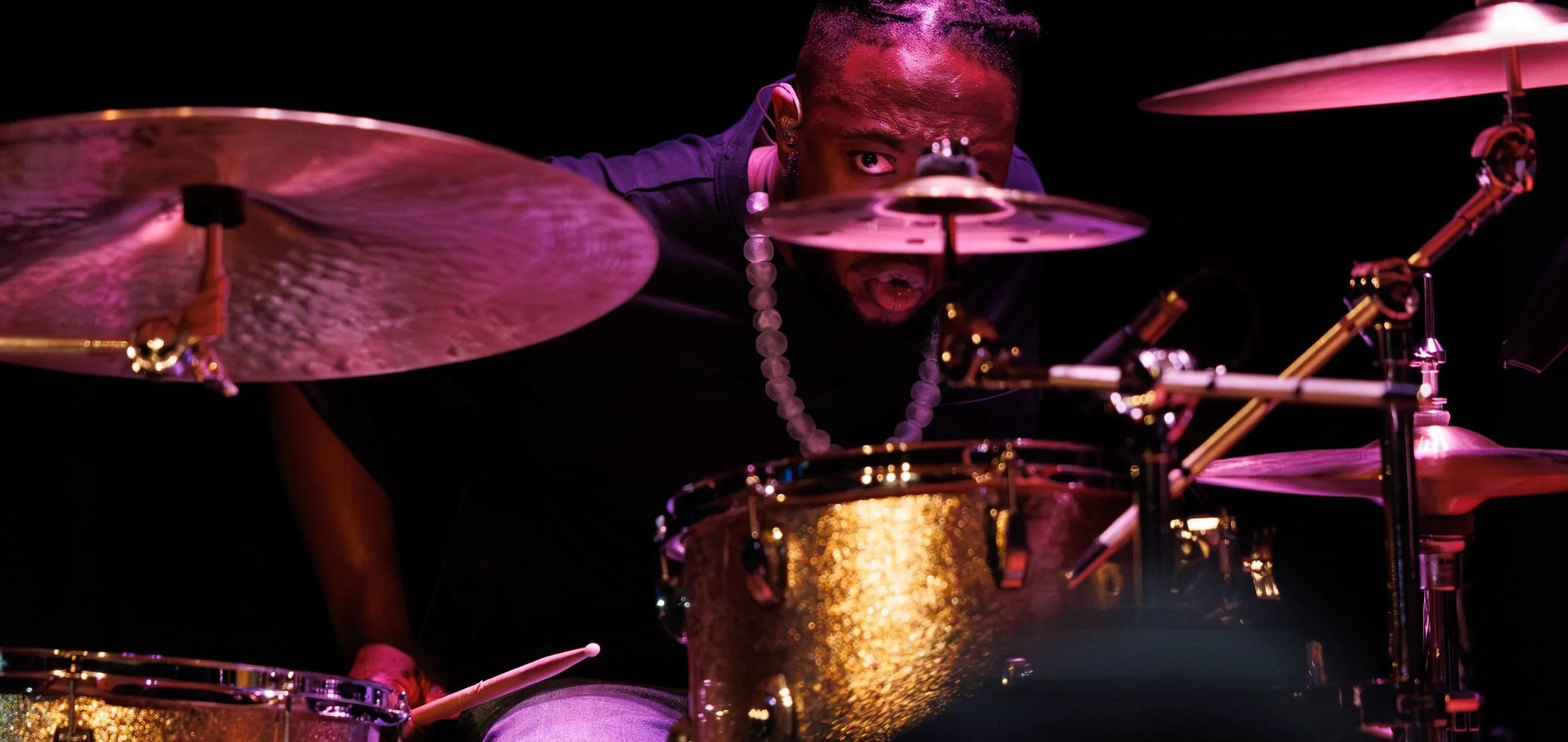 A drummer plays on stage, seen through the drum kit, with intense facial expression and focused eyes, illuminated by pink and purple stage lighting.