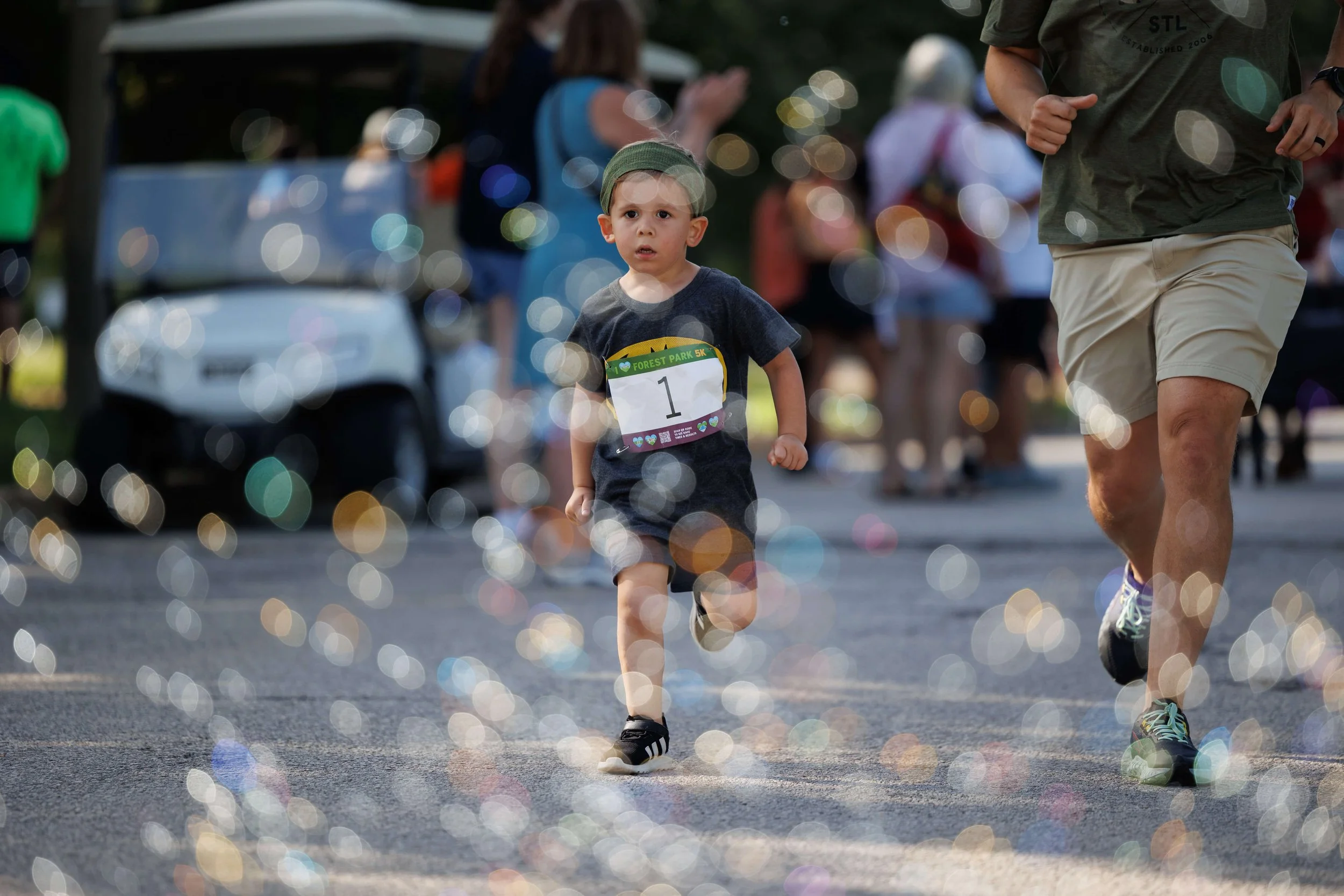 Young boy running through bubbles in a race with a bib number 1, surrounded by blurred spectators and a golf cart.
