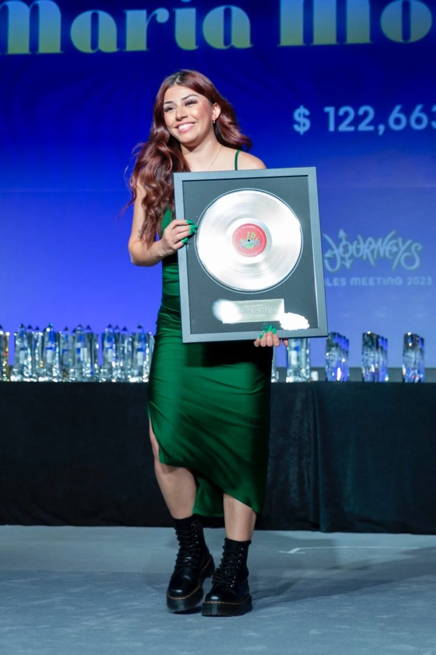 A woman in a green dress and black boots holding a platinum record award on stage at an event, with a showroom and awards in the background.