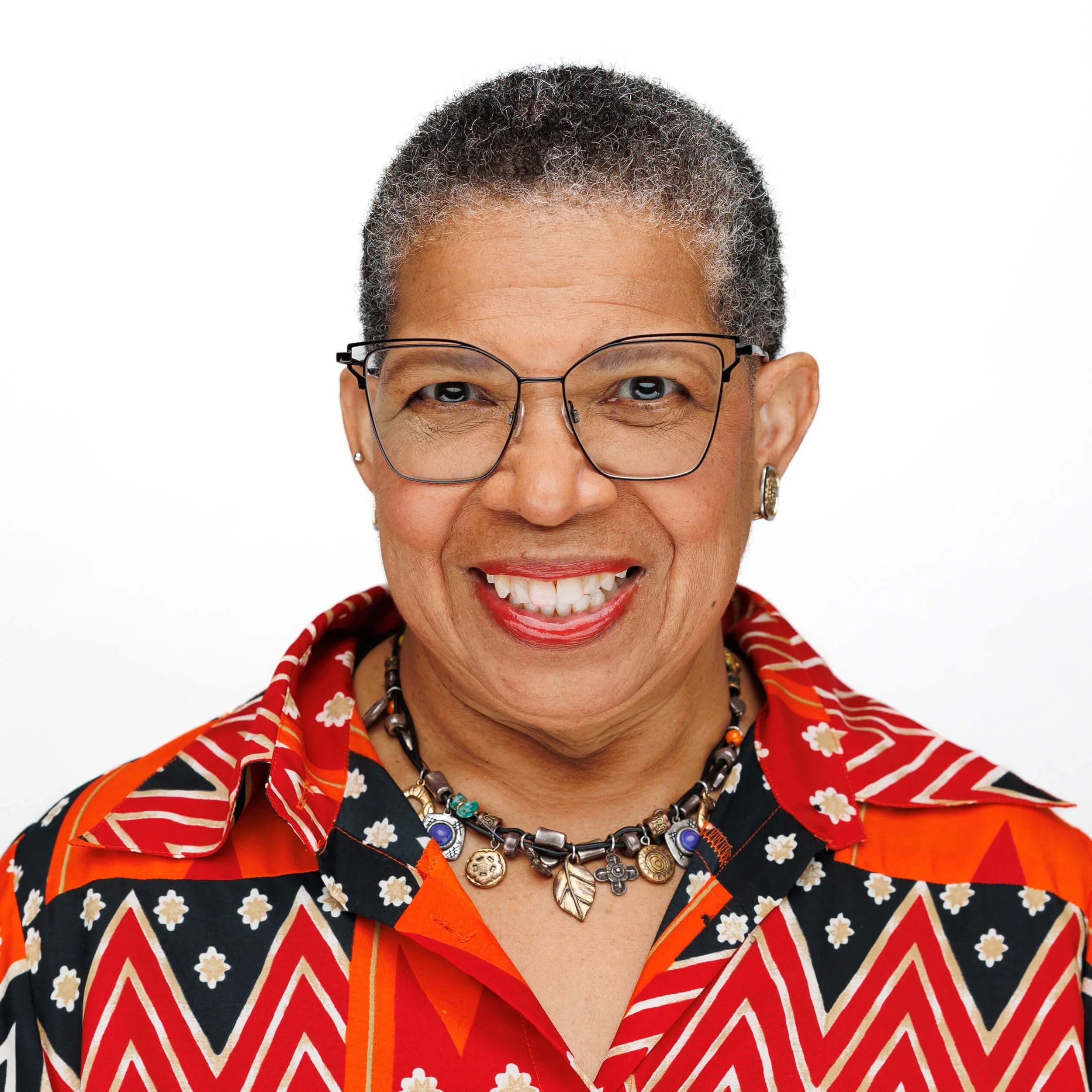 A smiling woman with short gray hair, wearing glasses, colorful patterned shirt, and layered necklaces.