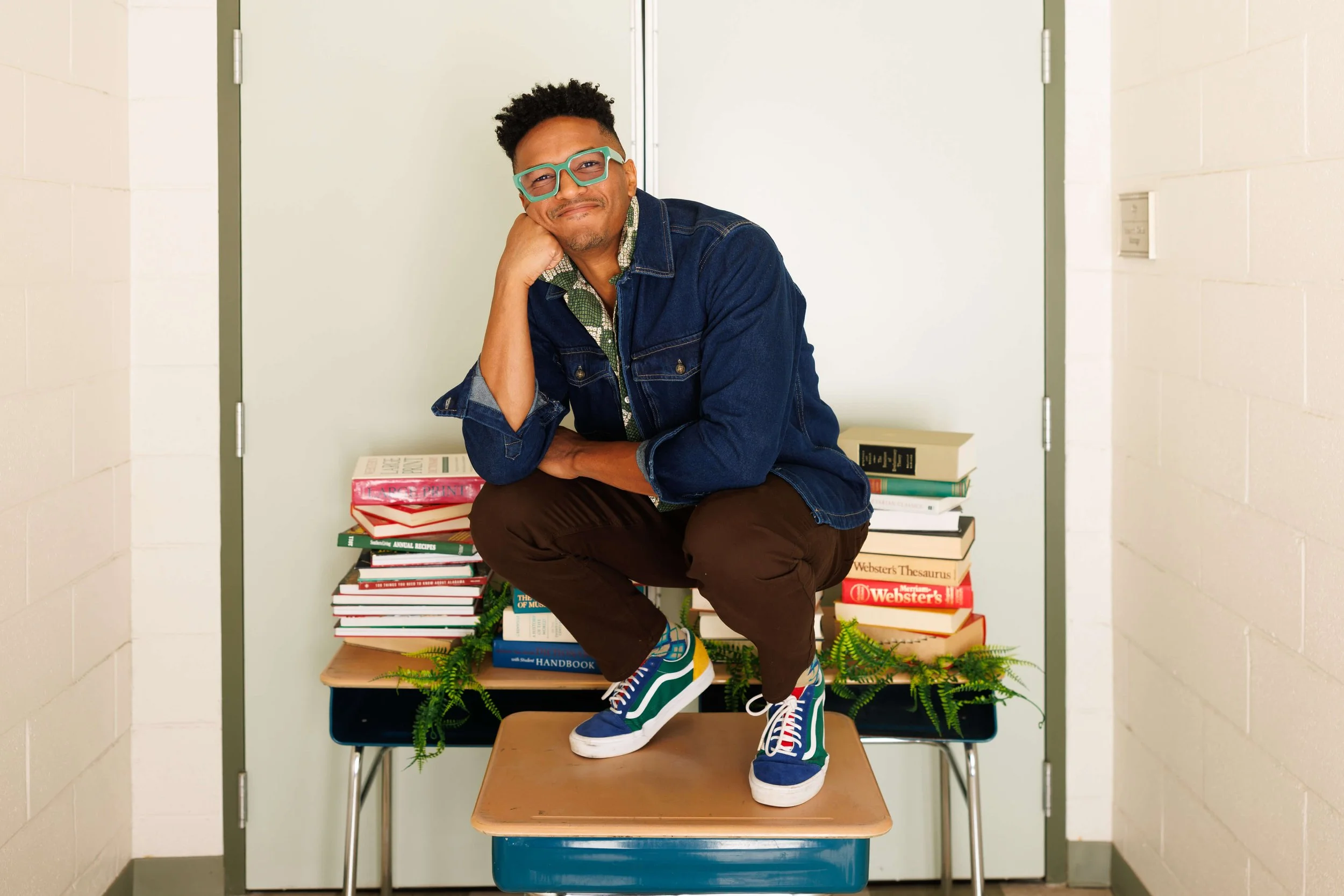 A man with glasses squatting on a desk covered with library books and green plants, making a playful face.