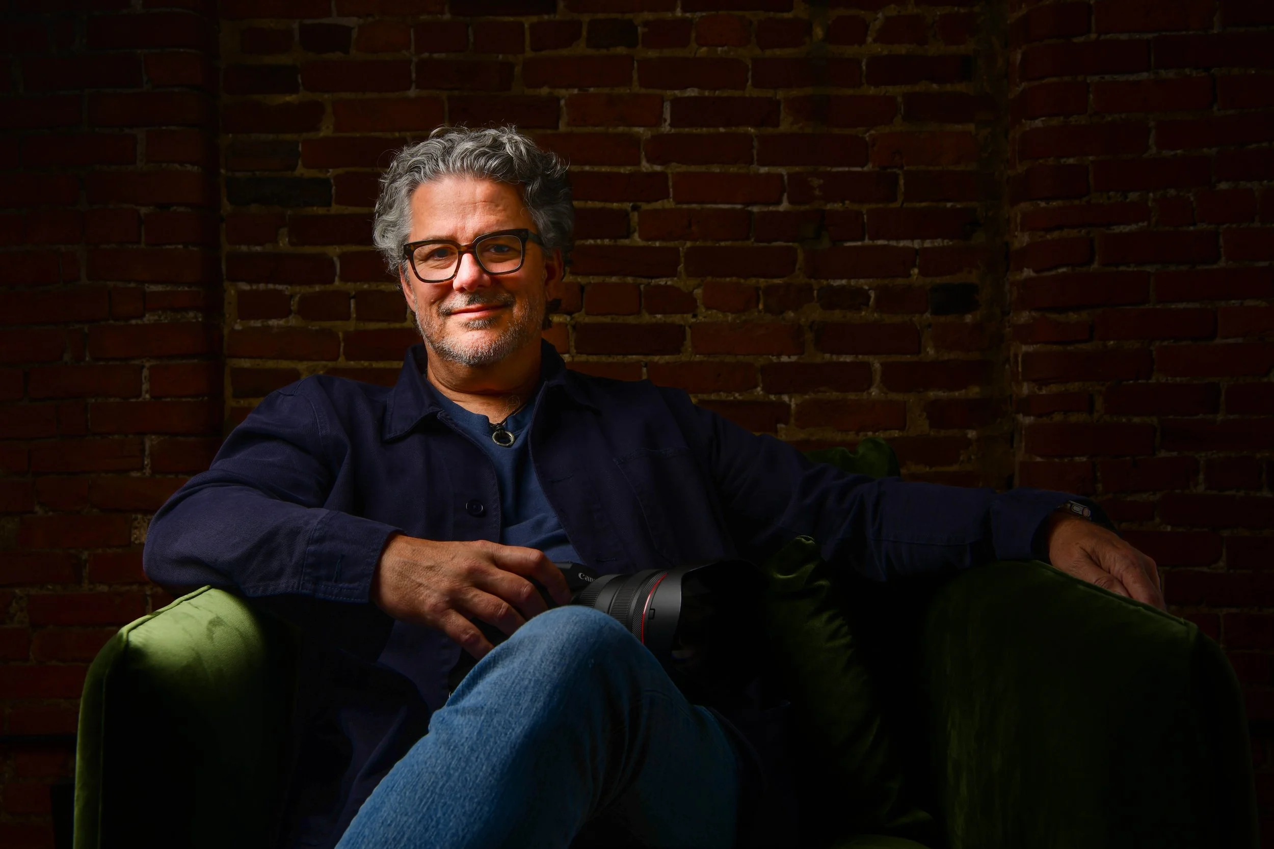 A middle-aged man with gray hair, glasses, and a beard sitting on a dark green sofa in front of a brick wall. He is holding a professional camera and smiling.