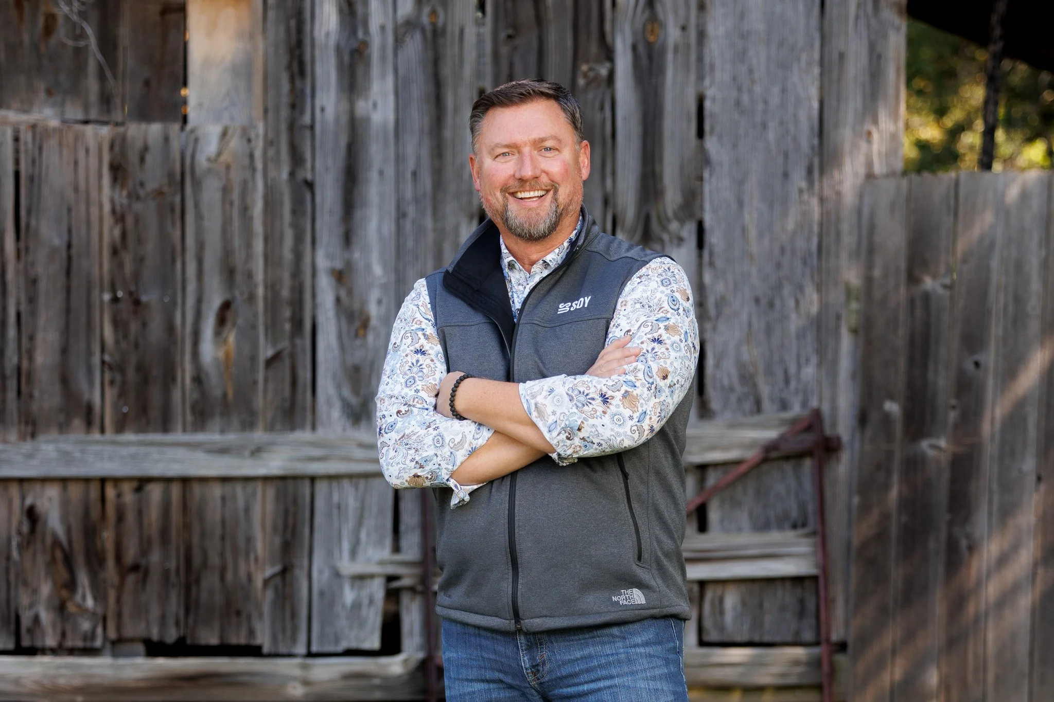 A smiling man with short brown hair and a beard standing outdoors in front of a wooden fence. He is wearing a patterned long-sleeve shirt, a gray sleeveless vest, and jeans, with his arms crossed.