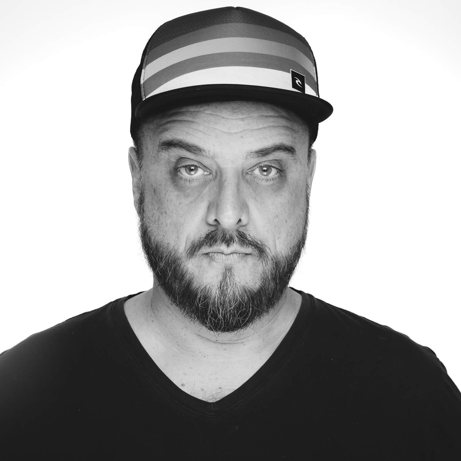 A black-and-white portrait of a man wearing a striped cap and a plain black shirt, with a beard and serious expression, looking directly at the camera.