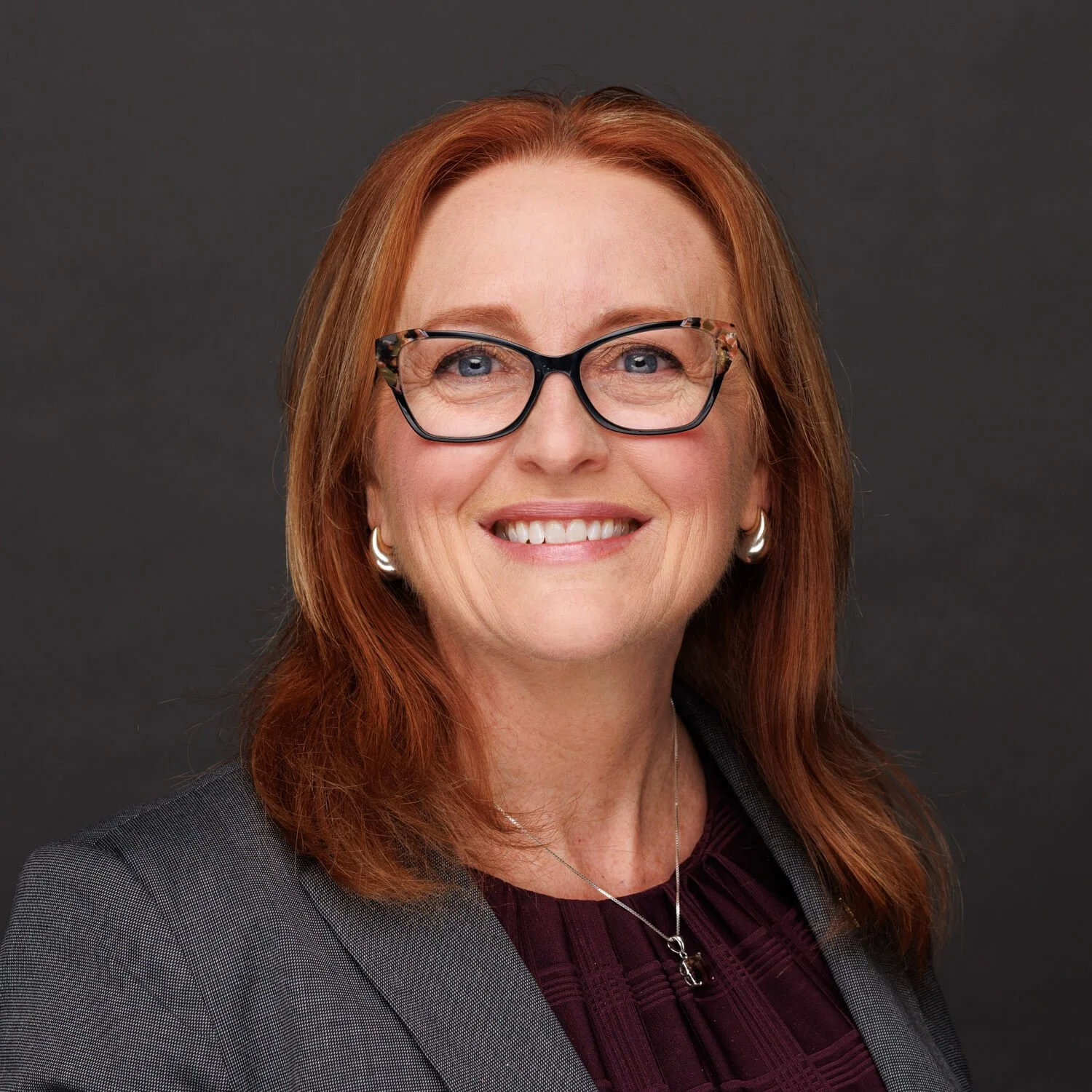 A portrait of a woman with red hair, wearing glasses, earrings, a necklace, a purple top, and a gray blazer, against a dark background.
