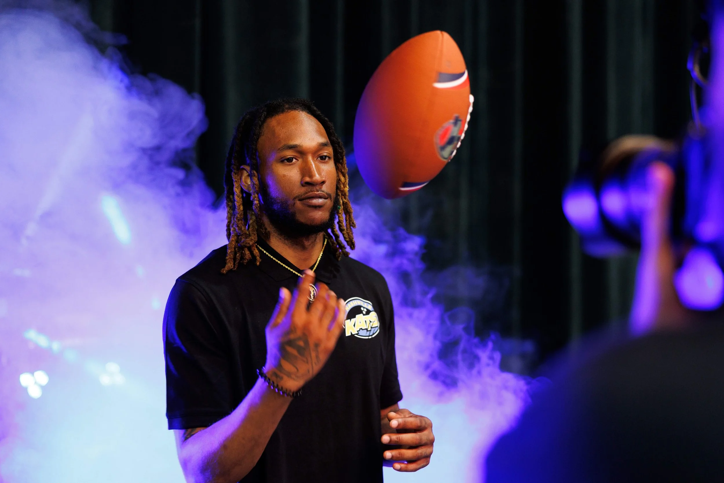 A man with dreadlocks stands in front of purple fog, wearing a black shirt with a sports team logo, and is about to catch a floating football.