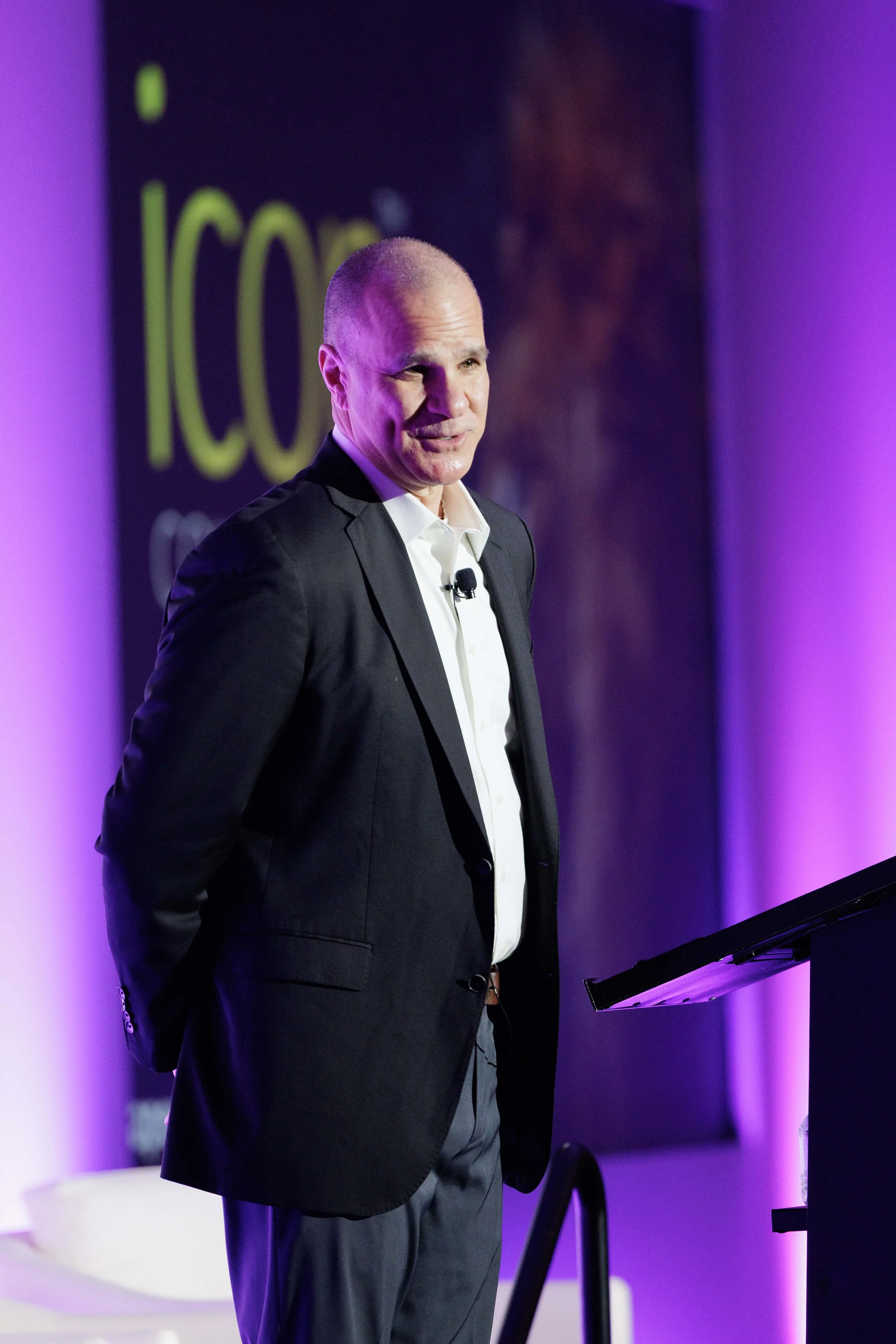 A man in a black suit and white shirt speaking at a conference with a purple-lit background.