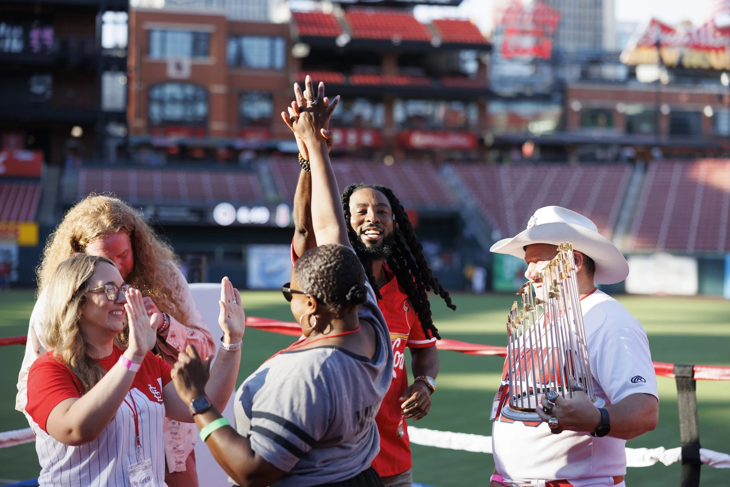 A group of people celebrating on a baseball field, with one person holding a trophy. The background shows stadium seats and a large digital scoreboard.